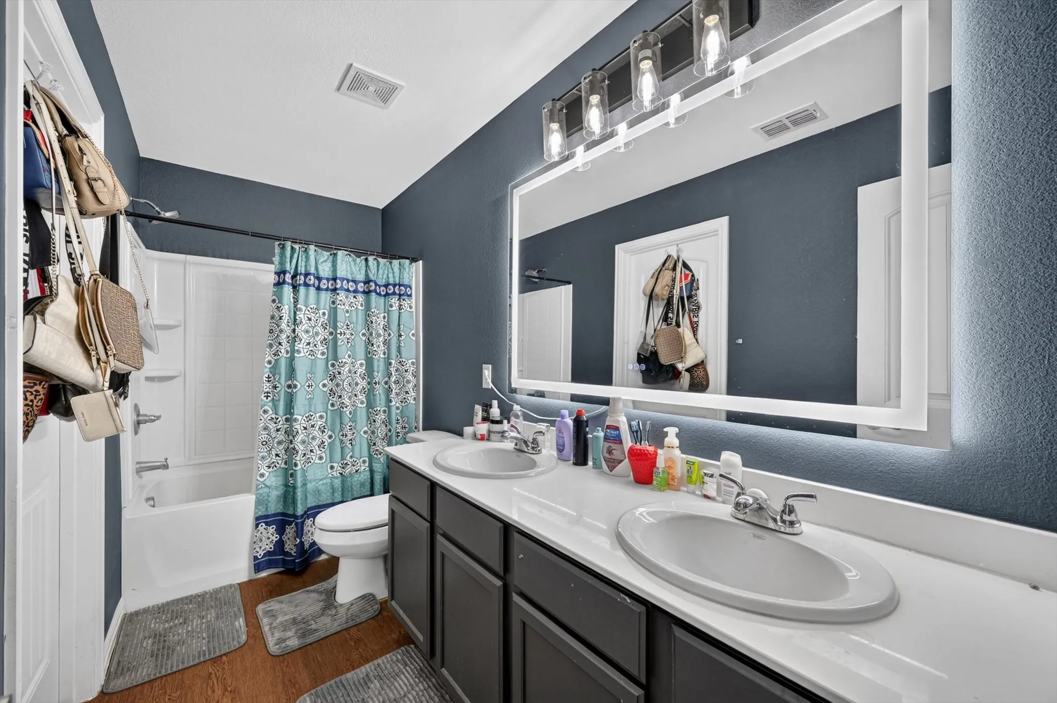Dual vanity with undermount sinks, illuminated mirror, wood-finish flooring, and a shower-tub combination