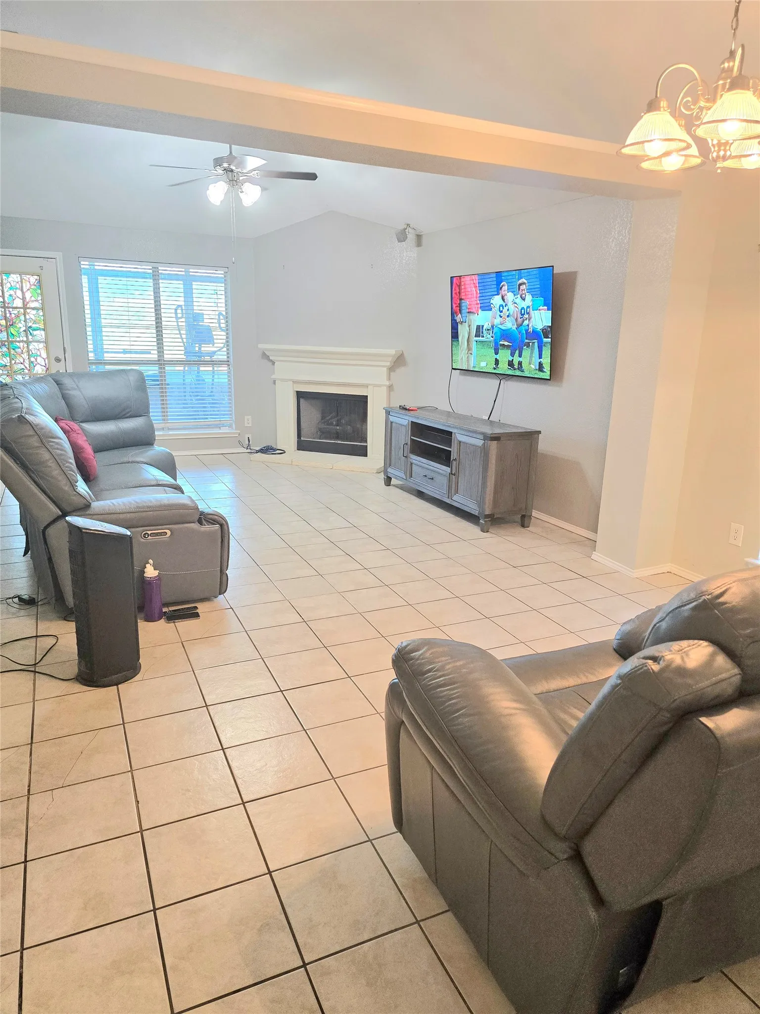 Vaulted ceiling with exposed beam, ceramic tile flooring, built-in fireplace with white mantel, ceiling fan, and large windows with blinds