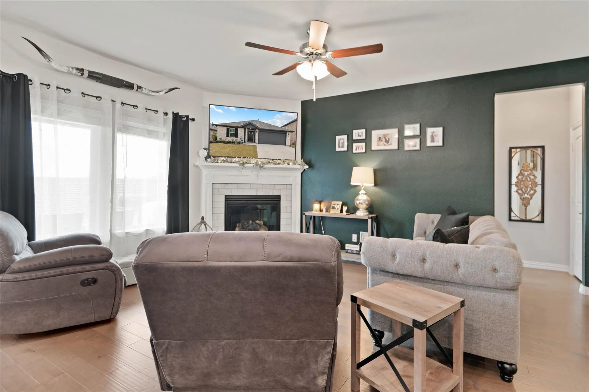 Living room featuring wood-finish flooring, a ceiling fan, a white brick fireplace with a mantel, and large windows