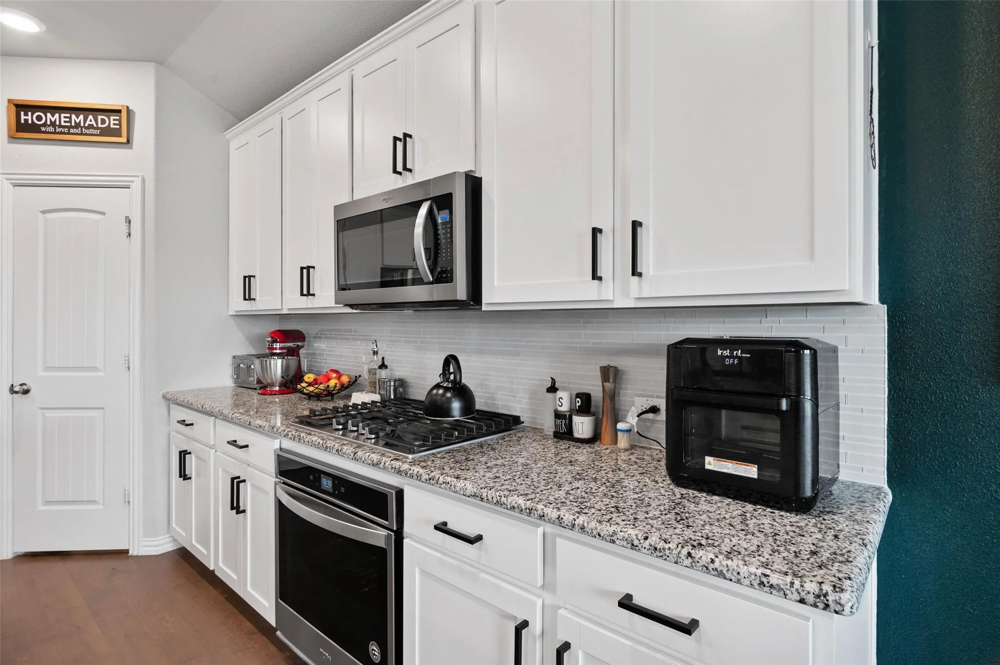 White shaker cabinetry with matte black hardware, granite countertops, stainless steel gas range with oven, built-in microwave, and tile backsplash