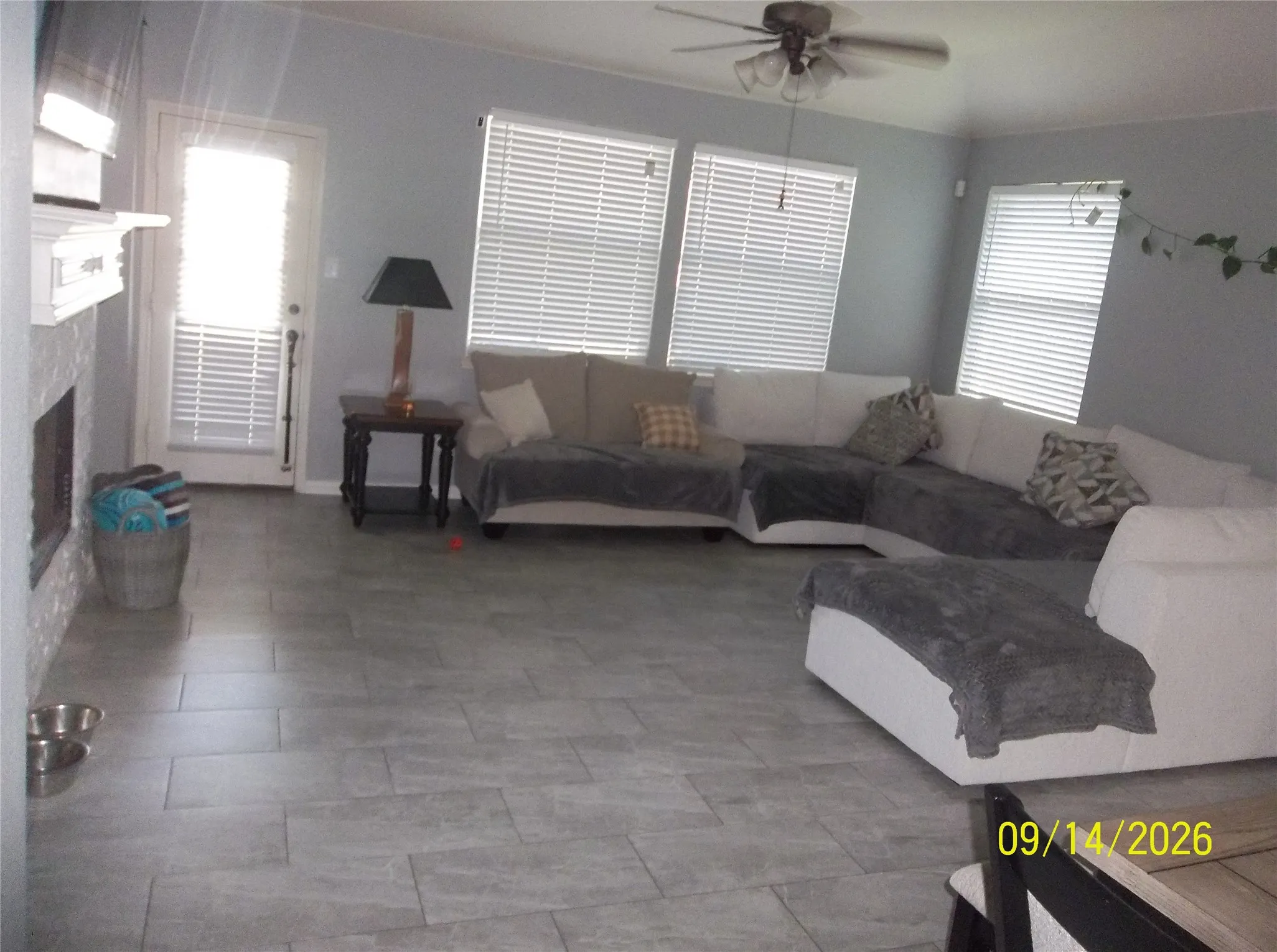 Spacious room featuring vaulted ceilings, a ceiling fan, multiple windows with blinds, a fireplace with a light-colored surround, and tile flooring