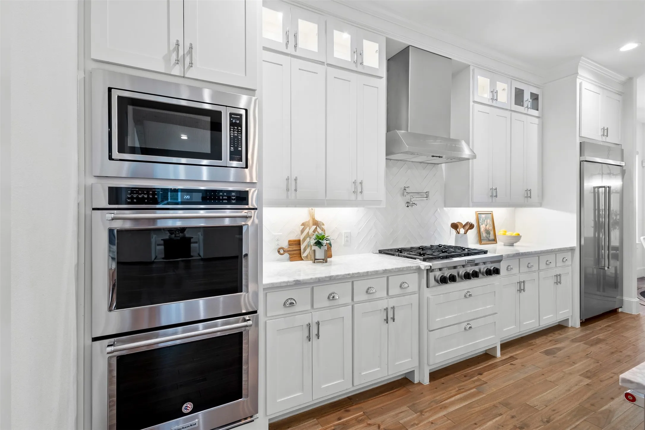White shaker cabinetry, stainless steel appliances, herringbone tile backsplash, light-toned countertops, wood-finish flooring