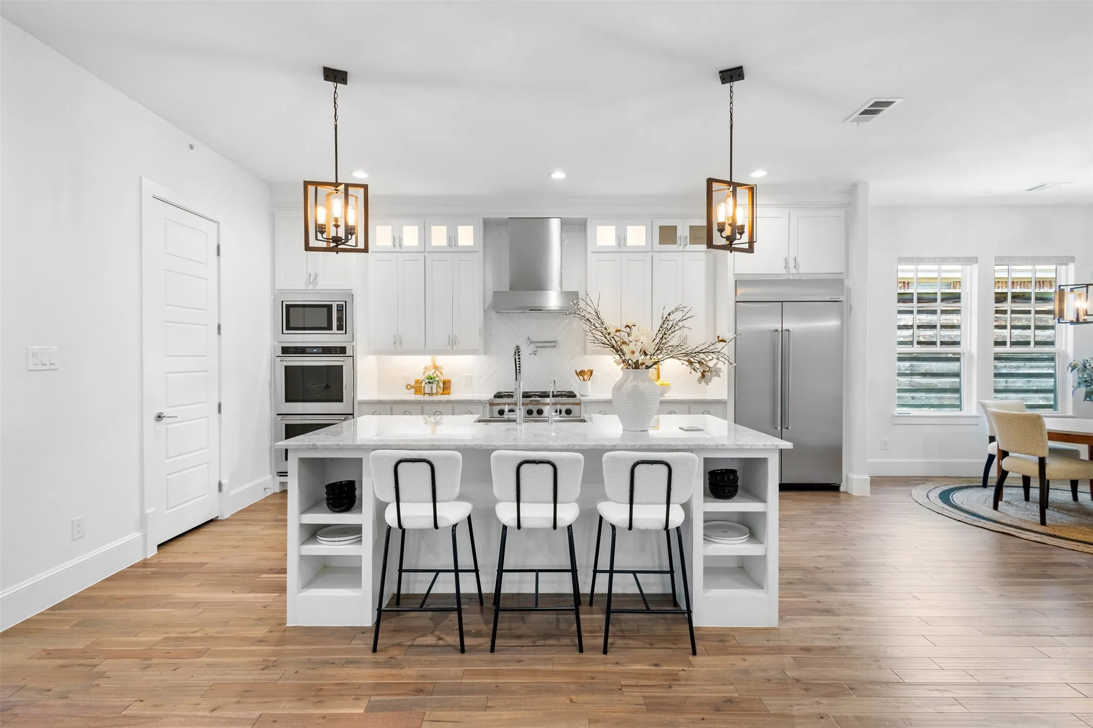 Open-concept kitchen featuring an oversized island with seating, stainless steel appliances, white cabinetry, wood-finish flooring, and two pendant light fixtures