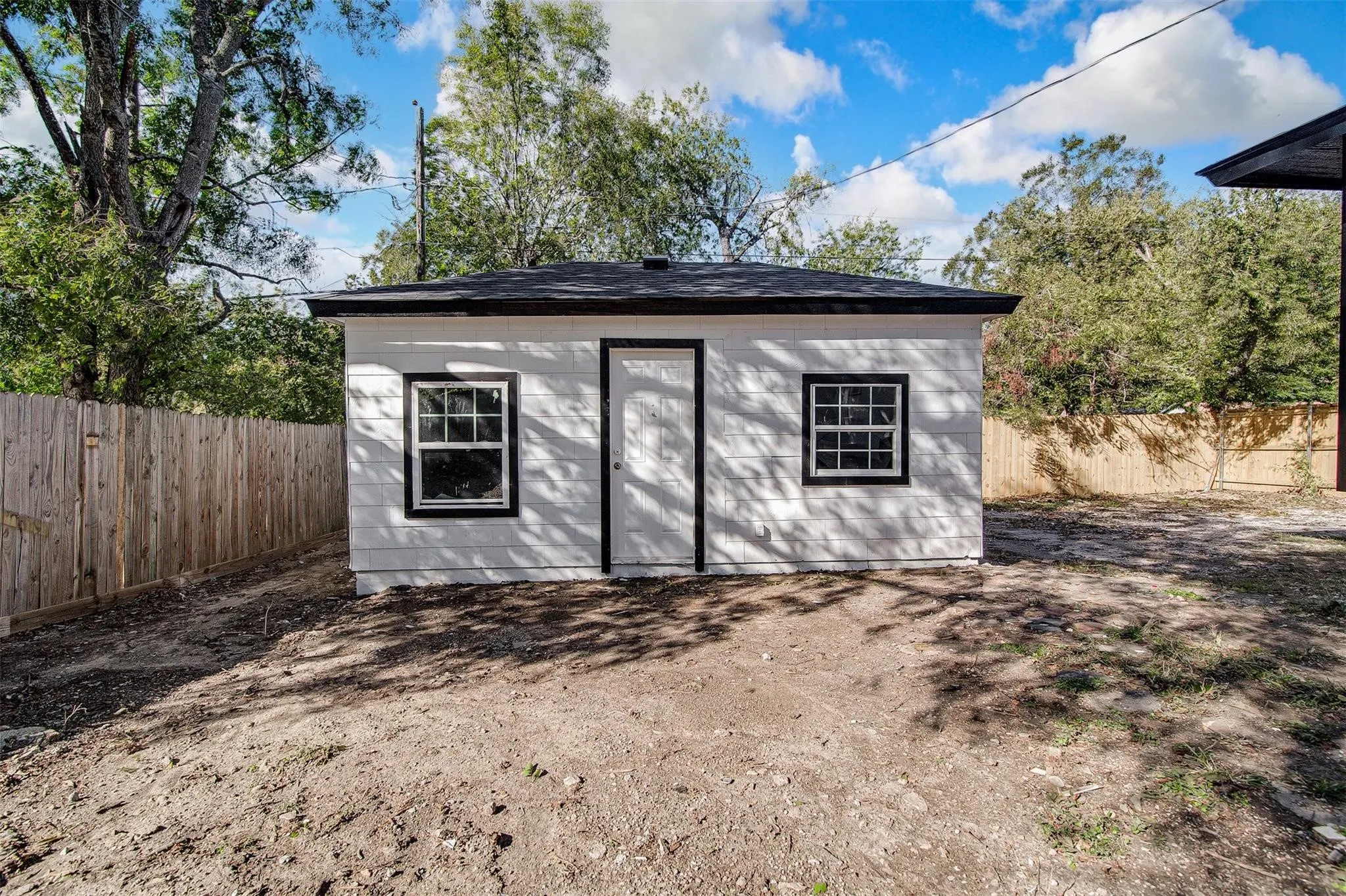Detached structure with white siding and a black shingle roof, featuring two single-hung windows with black trim and a central entry door