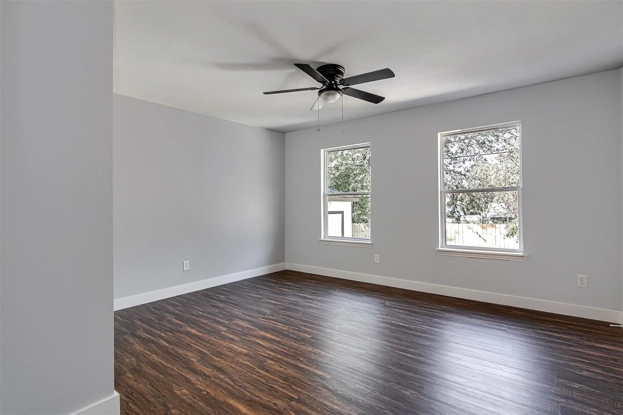 Spacious room featuring wood-finish flooring, light gray walls, two windows, and a ceiling fan with integrated lighting