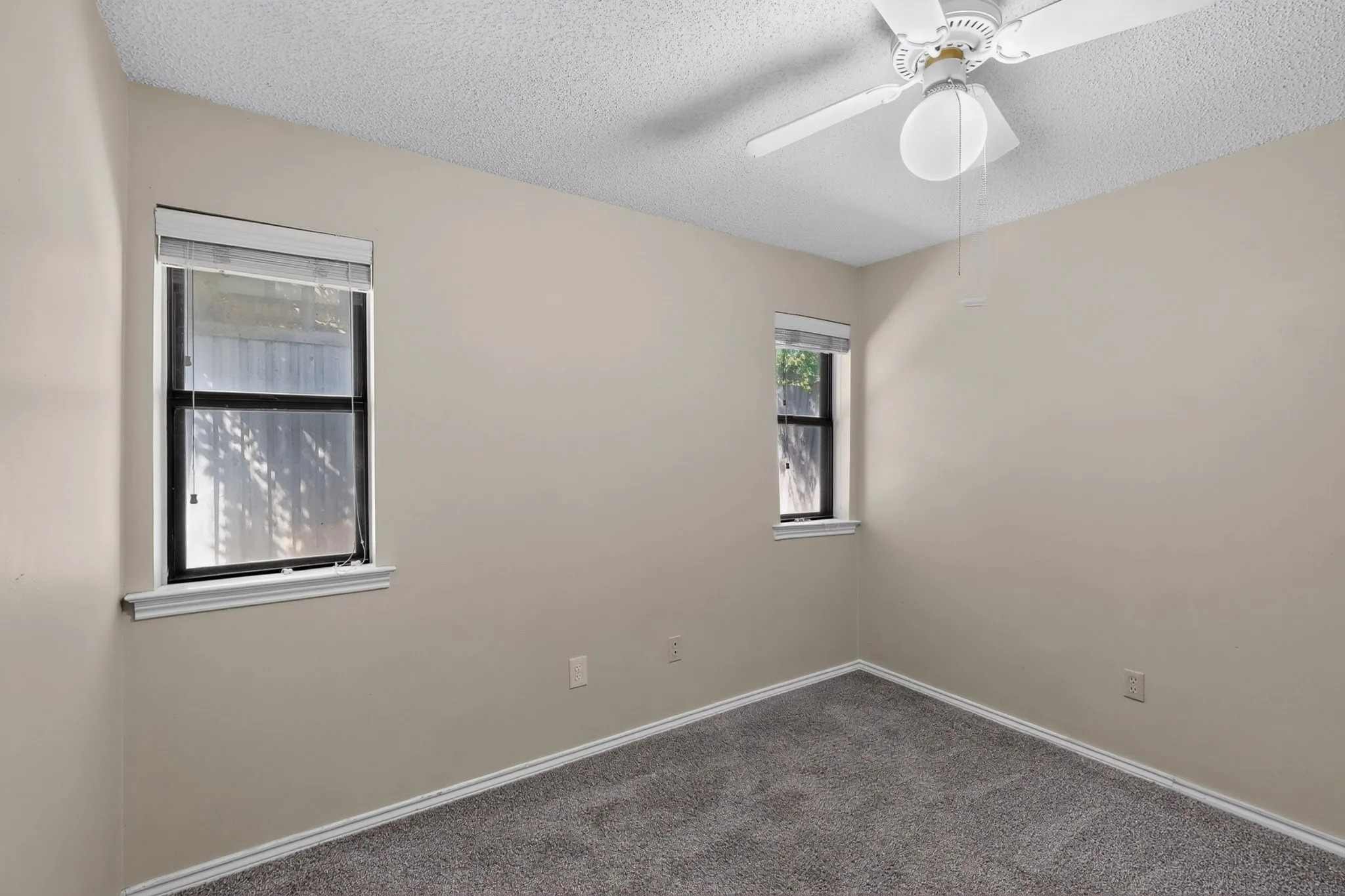 Textured ceiling with a ceiling fan light fixture, two windows with blinds, and gray carpeting