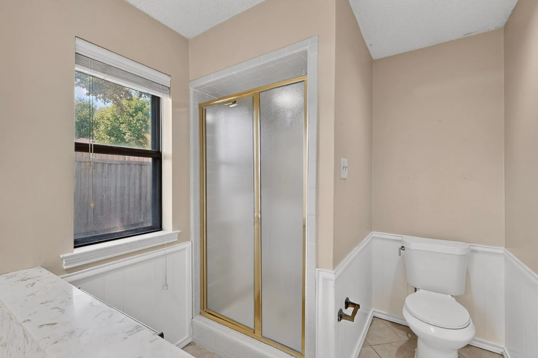 Shower with frosted glass doors and gold-tone framing, white wainscoting, and a window with blinds