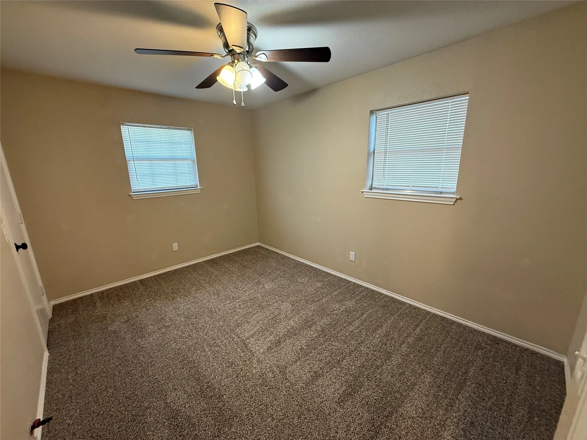 Neutral tone wall paint, textured carpet flooring, two windows with blinds, and a ceiling fan with integrated lighting