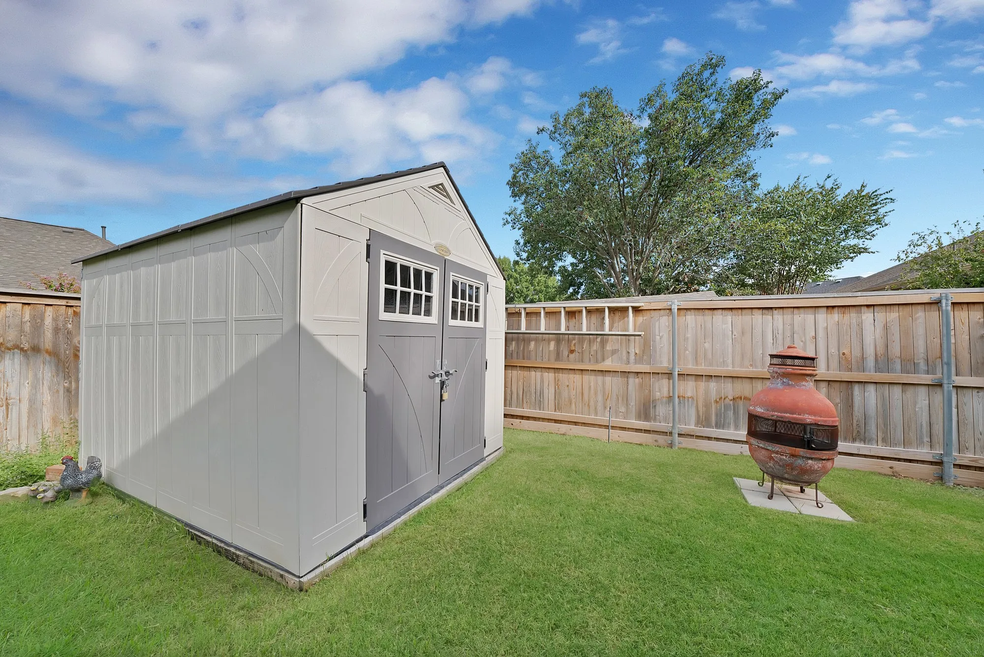 Storage shed with double doors and window accents, set on a manicured lawn, bordered by a wood privacy fence