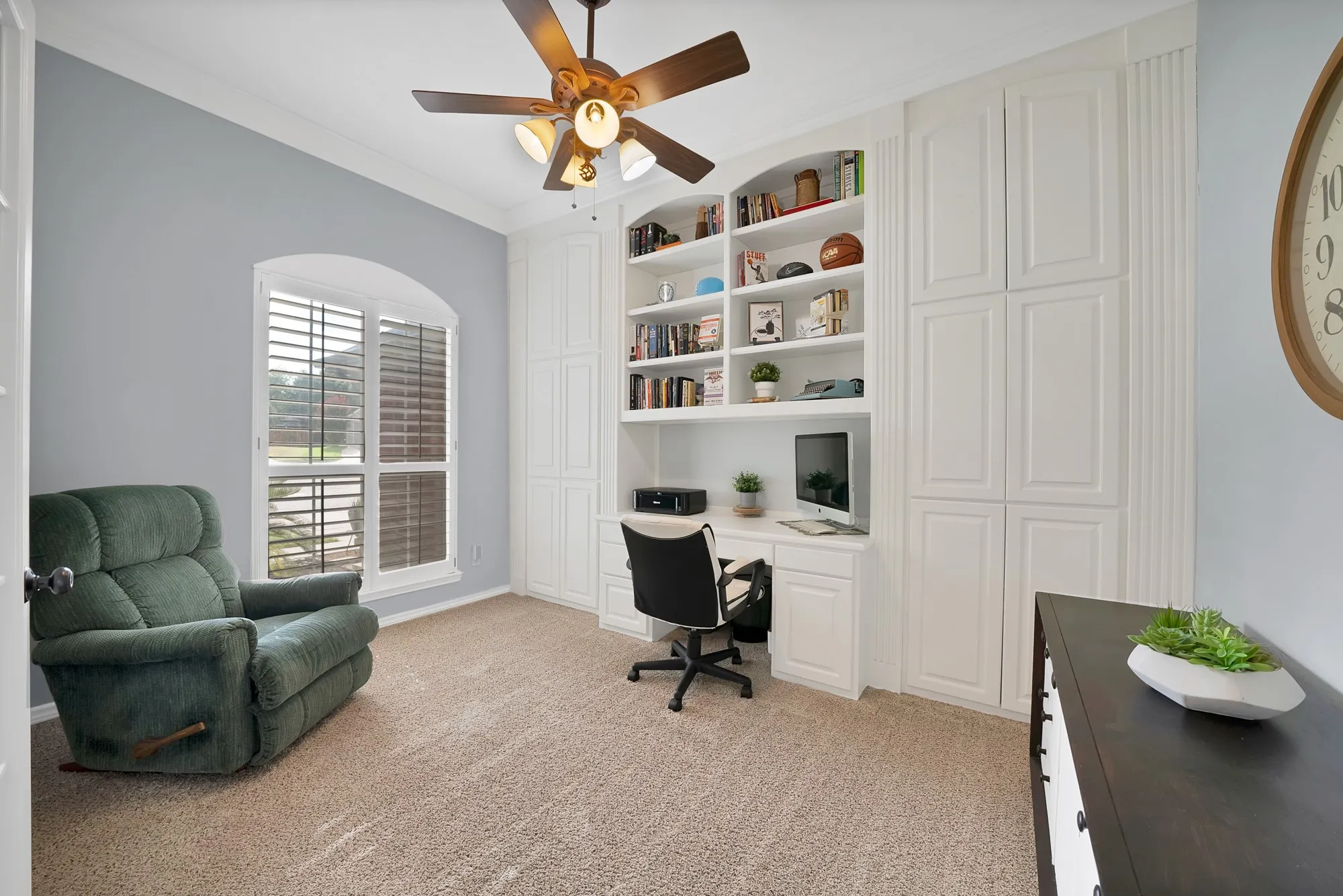 Built-in shelving and cabinetry with an integrated desk, arched window with plantation shutters, ceiling fan, and carpeting