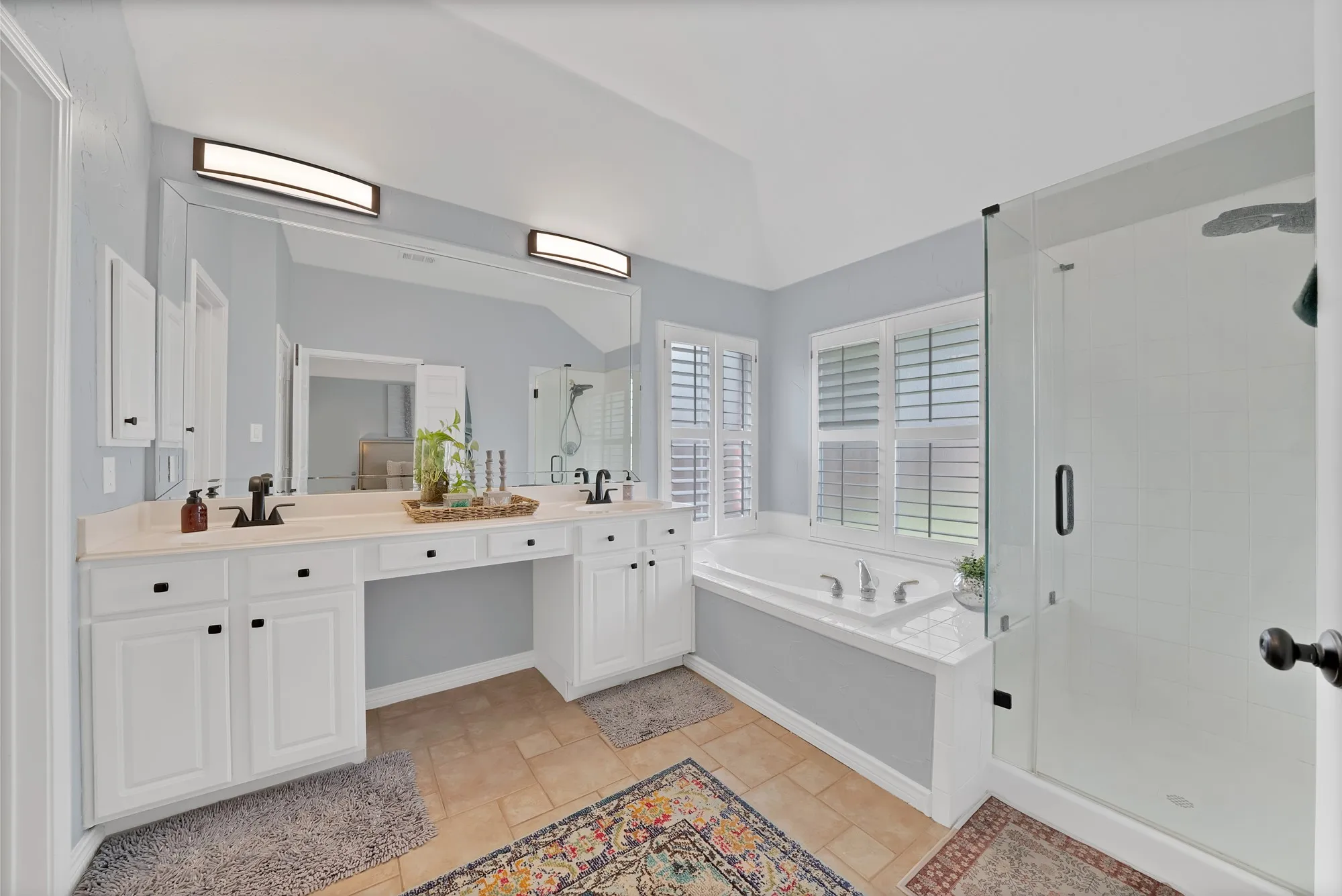Dual vanities with white cabinetry, built-in soaking tub with white surround, glass-enclosed shower, and plantation shutters