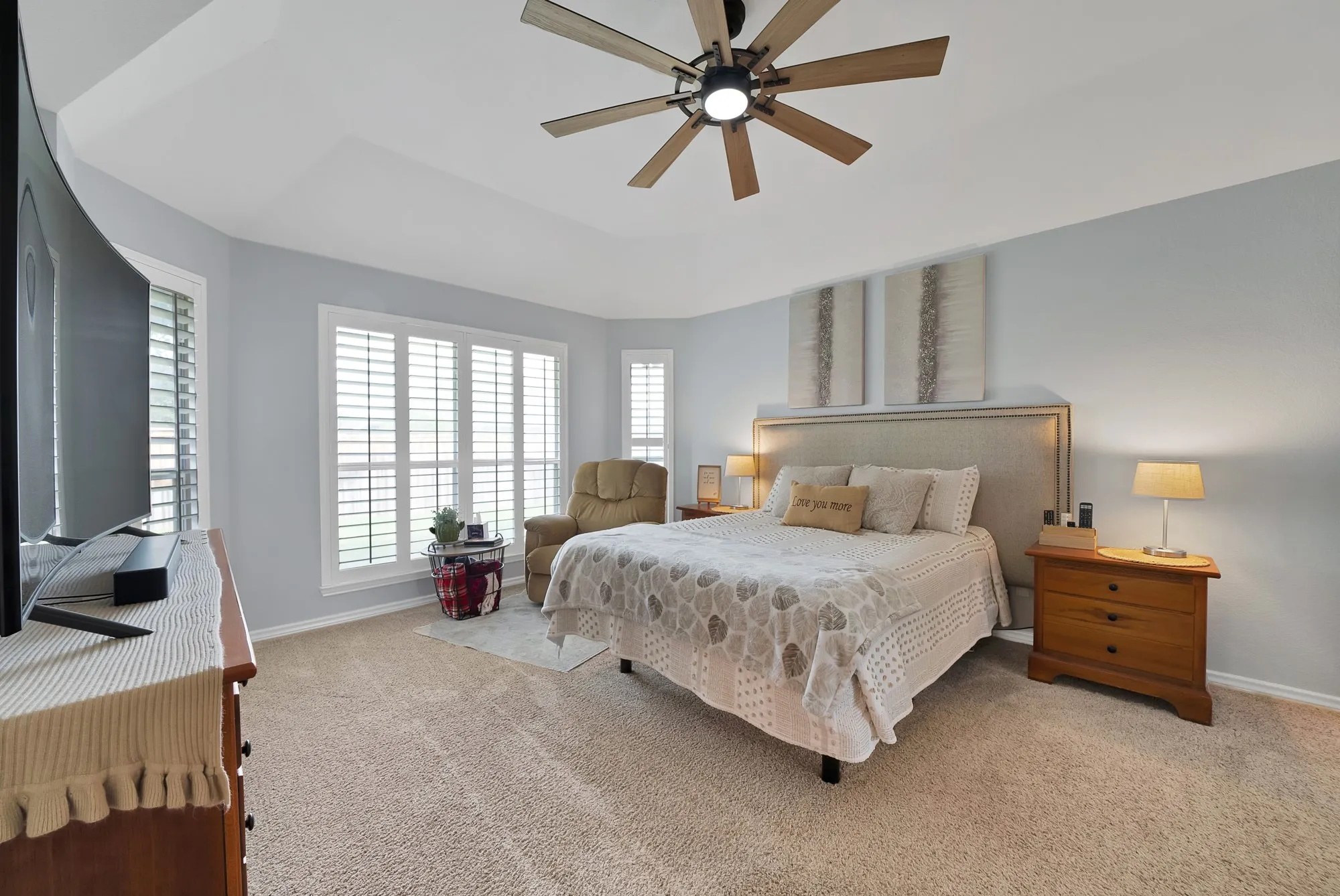 Tray ceiling with a ceiling fan, light-colored carpeting, multiple windows with white plantation shutters, and a fabric-upholstered headboard