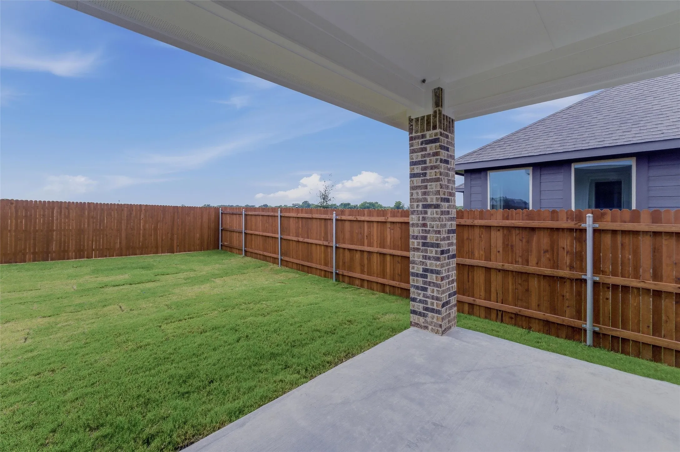 Expansive backyard featuring a concrete patio, brick support column, wood privacy fencing, and green lawn space