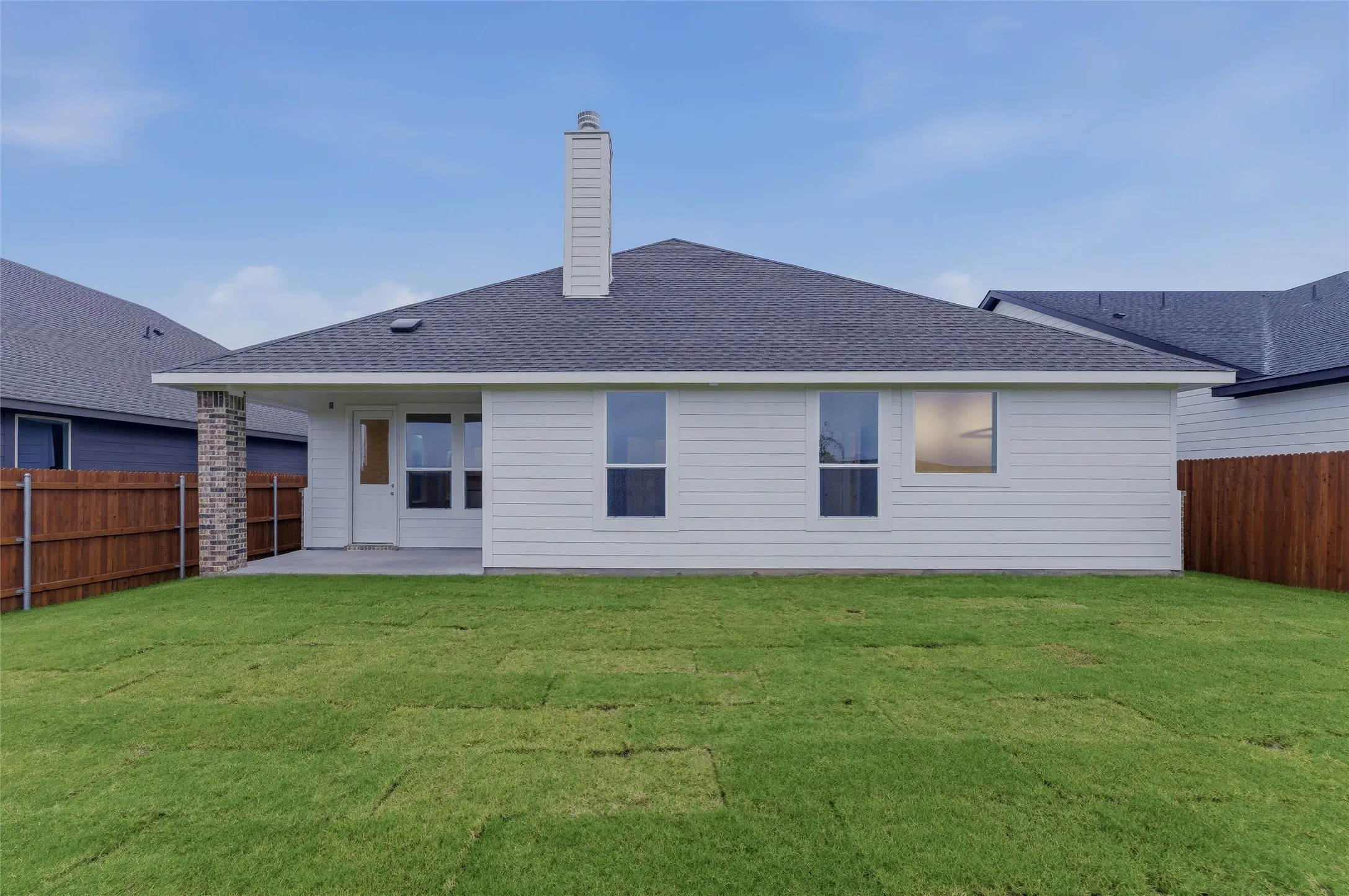 White siding exterior featuring a covered patio, brick accent column, a single chimney, and a dark shingle roof