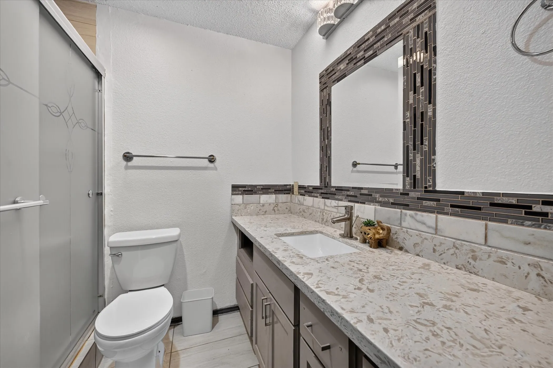 Frosted glass shower enclosure, single vanity with integrated sink, stone-look countertop, tile backsplash, and mirror with tile frame