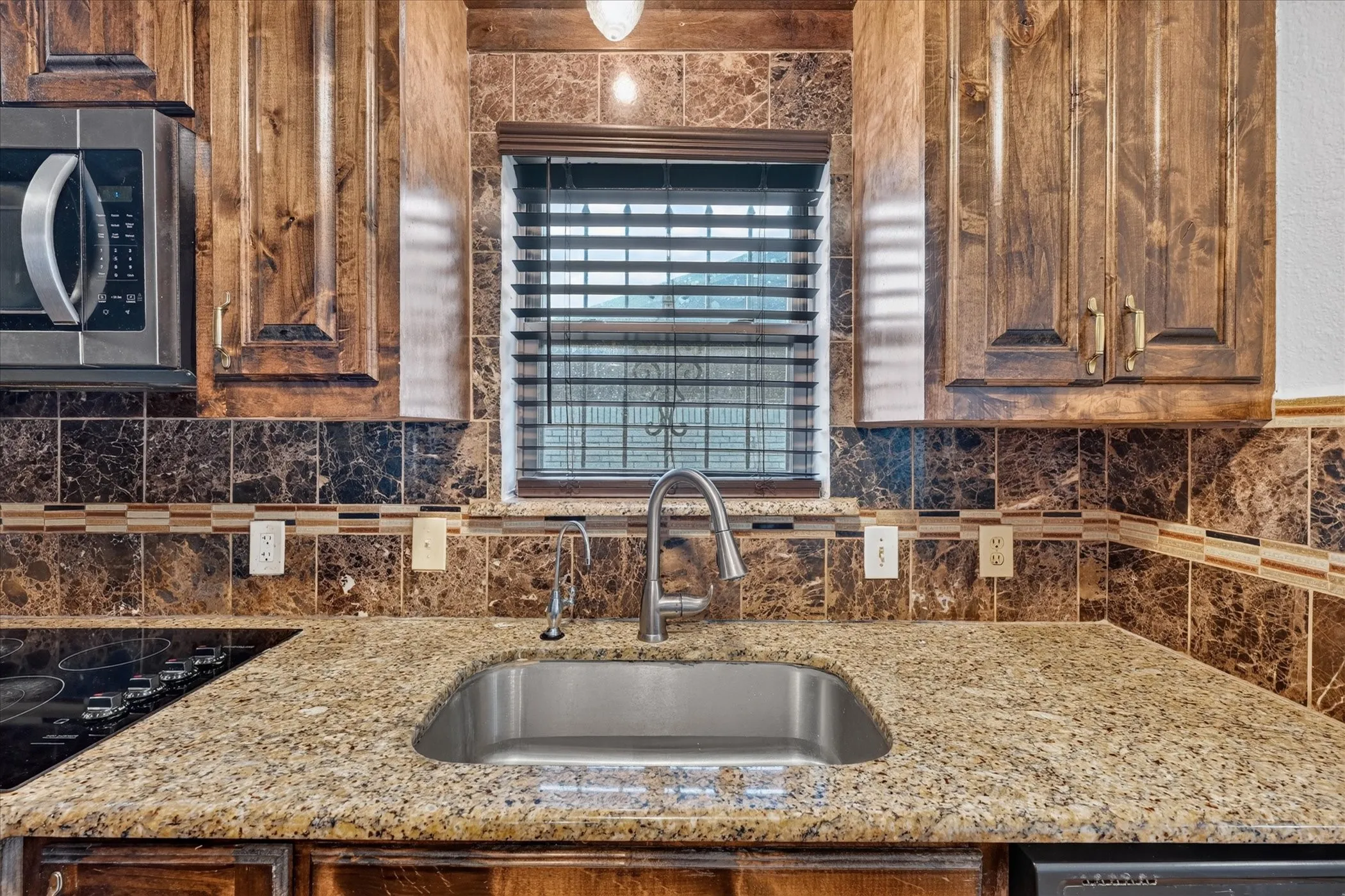 Granite countertops, stainless steel sink, wood cabinetry, built-in microwave, and tile backsplash with decorative accent banding