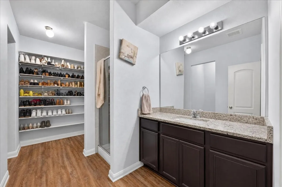 Wood-finish flooring throughout, vanity with a stone countertop and dark cabinetry, large mirror with overhead lighting, glass-enclosed shower, and a dedicated closet space with built-in shelving