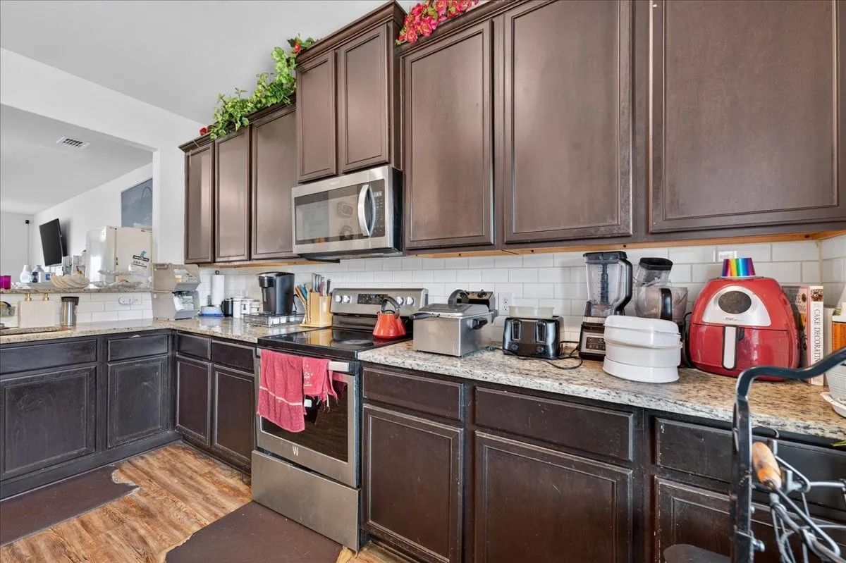 Kitchen featuring dark wood-finish cabinetry, light-colored speckled countertops, stainless steel appliances, a tile backsplash, and wood-finish flooring