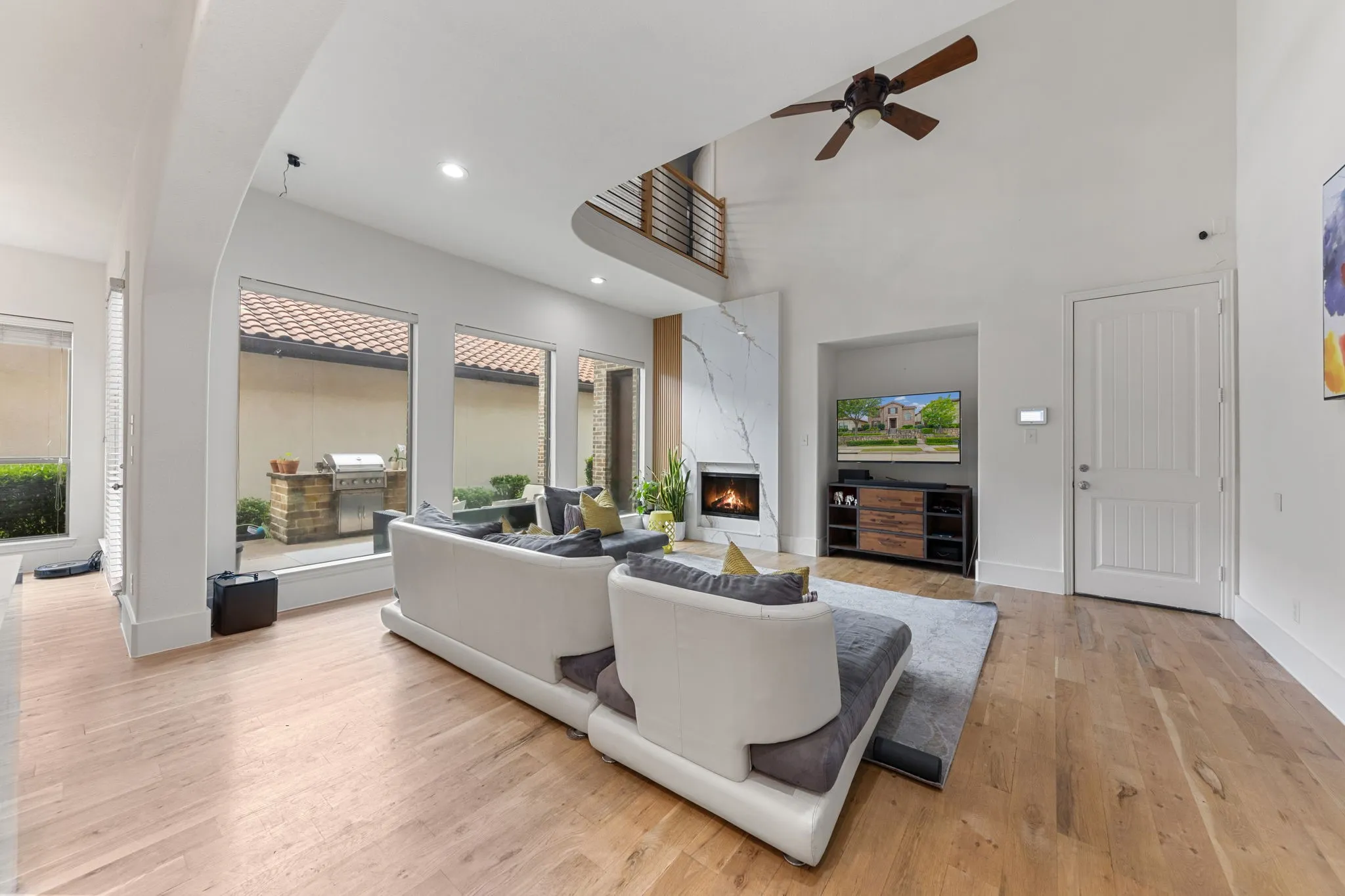Vaulted ceiling with a ceiling fan, fireplace with a stone-finish facade, wood-finish flooring, built-in media niche, and an exterior patio with a built-in grill