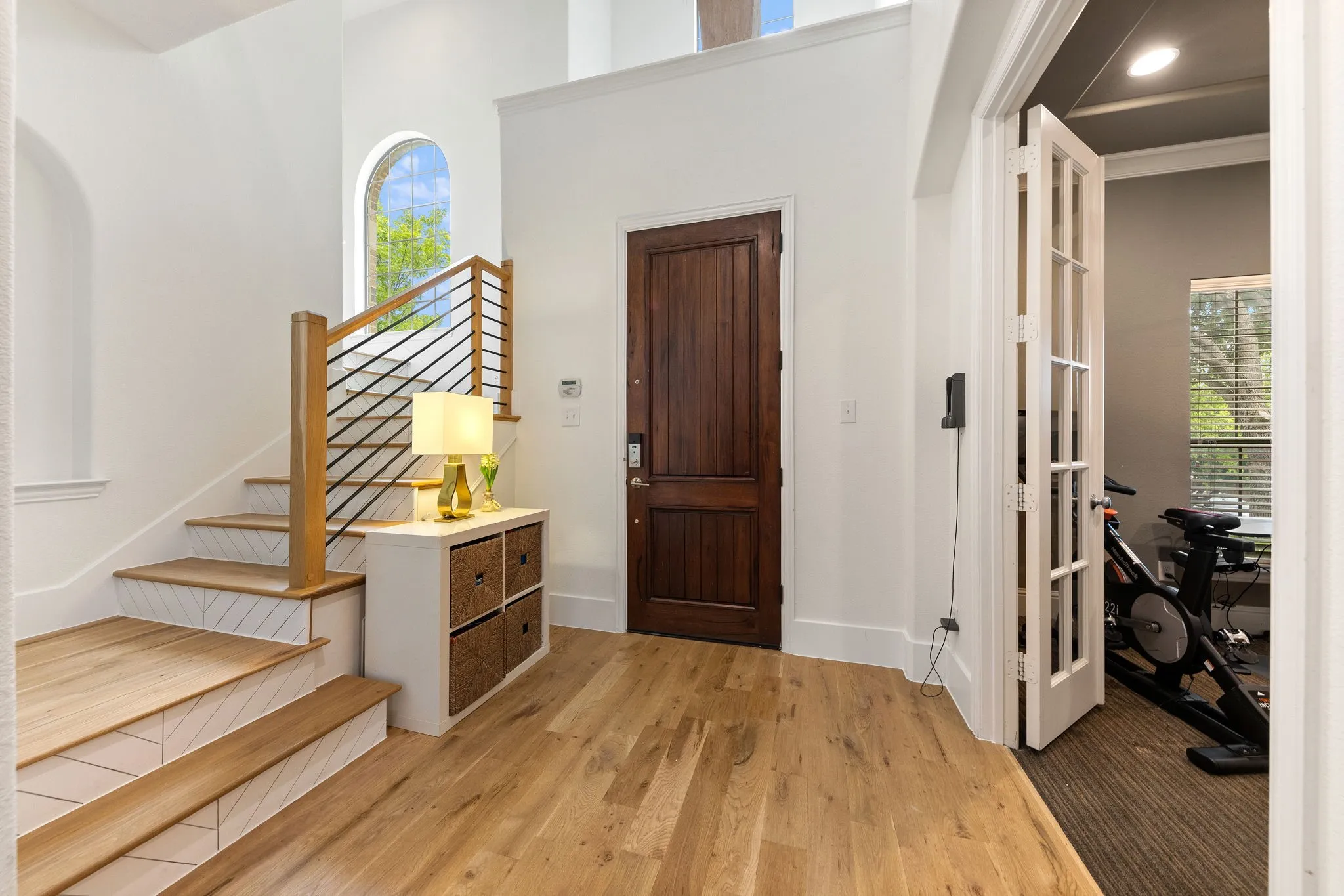 Light wood-finish flooring, dark wood entry door, staircase with wood treads and metal balusters, arched window, and French doors with glass panels