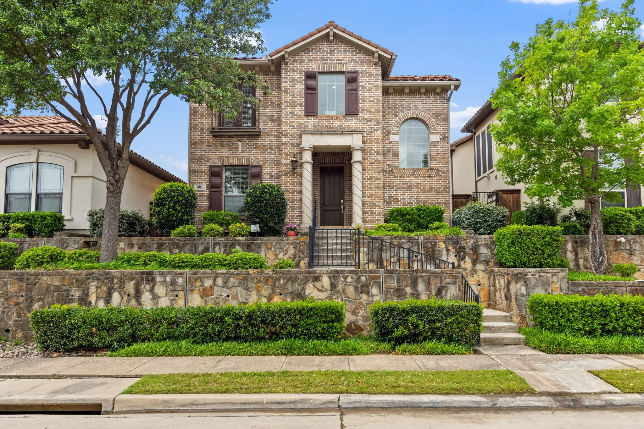 Brick facade with stone accents, arched windows, covered entry, and tiered landscaping with mature trees and shrubs