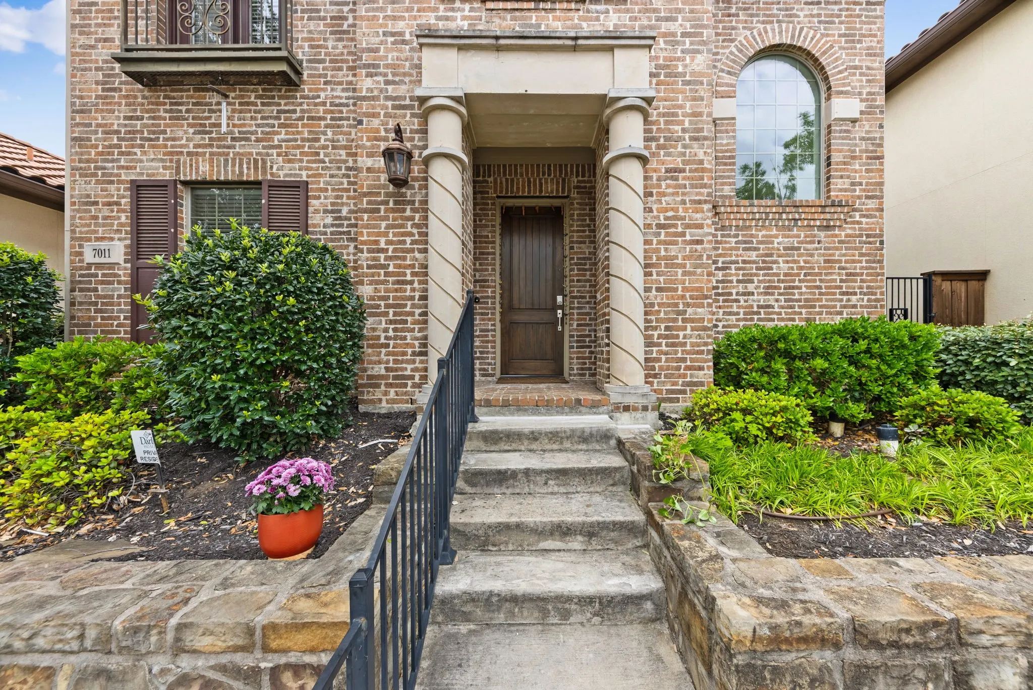 Brick exterior with stone accents, arched window, wrought iron railing, and mature landscaping