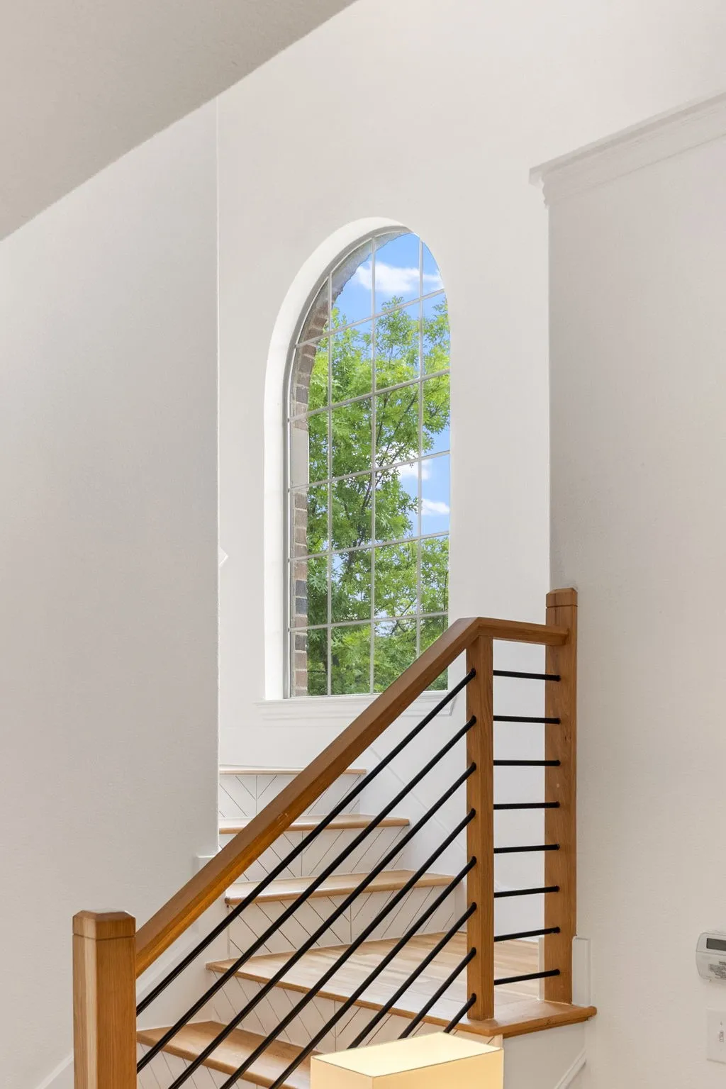 Arch-top window with grid detailing, natural wood staircase railing, horizontal metal balusters, and crown molding