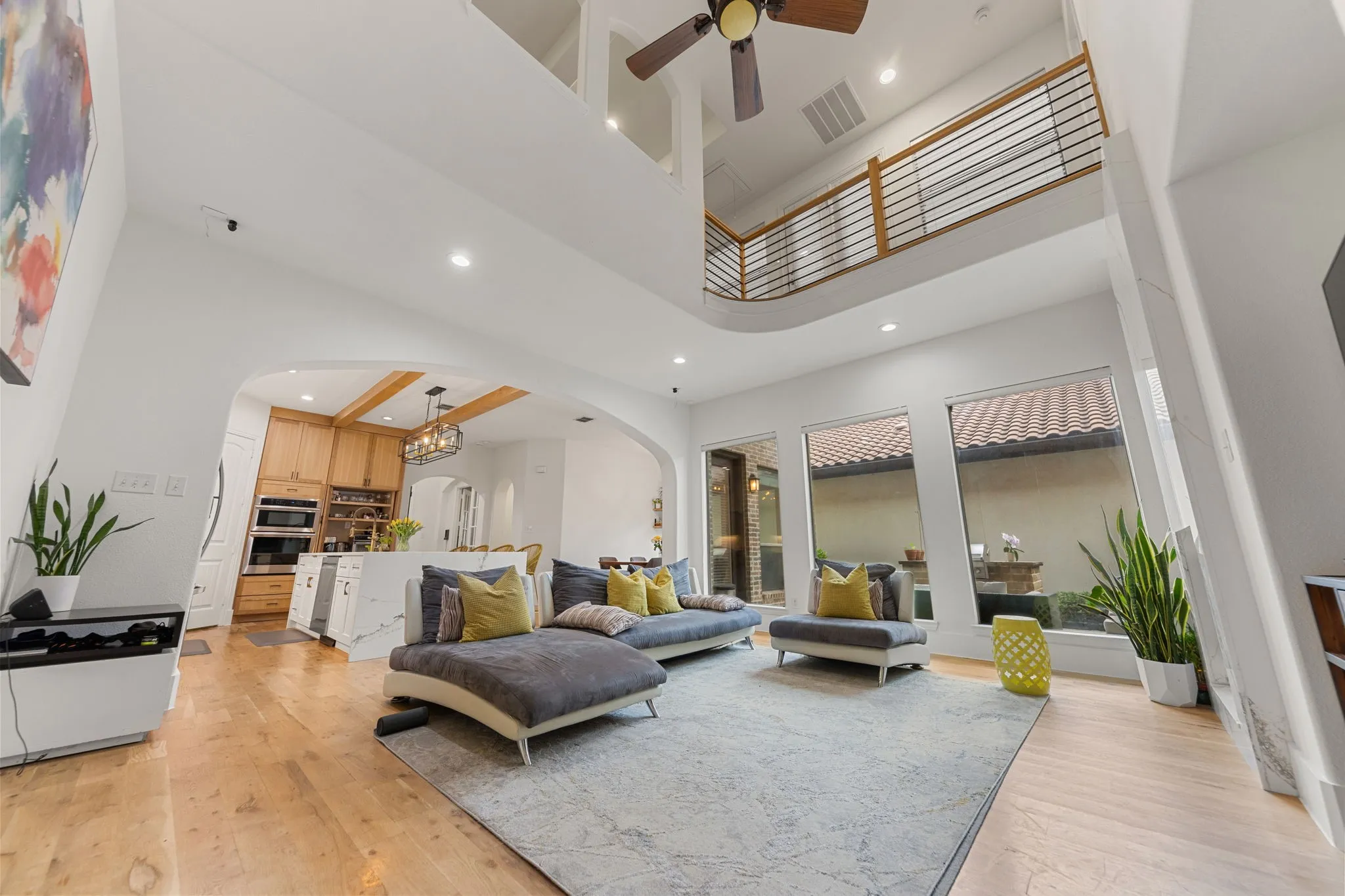 Double-height ceiling with recessed lighting, wood-finish flooring, arched entryway to the kitchen, a curved balcony with metal railings, and multiple windows
