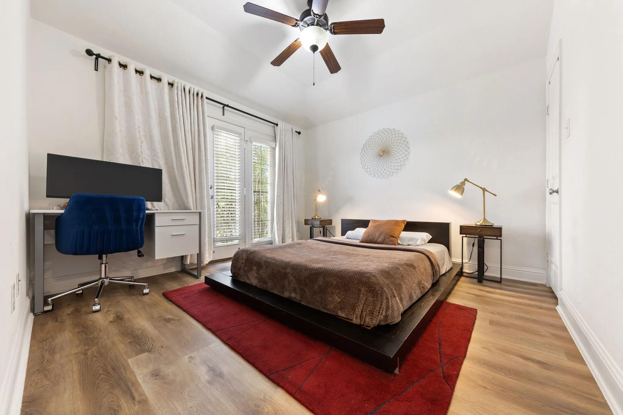 Spacious bedroom featuring hardwood flooring, a ceiling fan with integrated lighting, and large windows with plantation shutters and layered curtains providing natural light and privacy