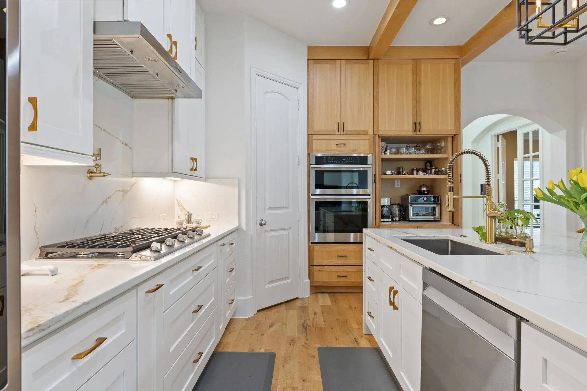 White shaker cabinetry with gold-tone hardware, stainless steel gas cooktop, double wall ovens, wood-finish flooring, and a kitchen island with an undermount sink