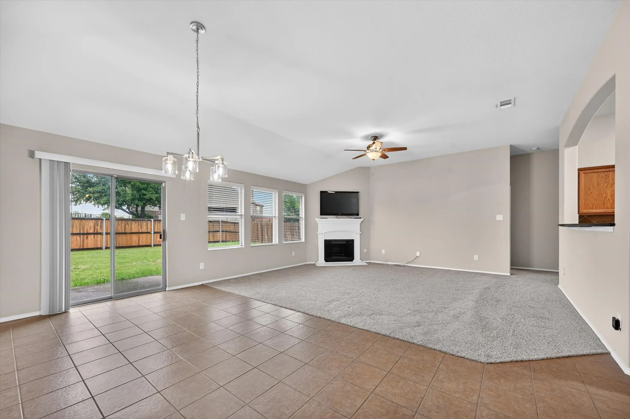 Vaulted ceiling with a contemporary chandelier and ceiling fan, tiled flooring, carpeted living area, fireplace with white mantel, and sliding glass door to the backyard