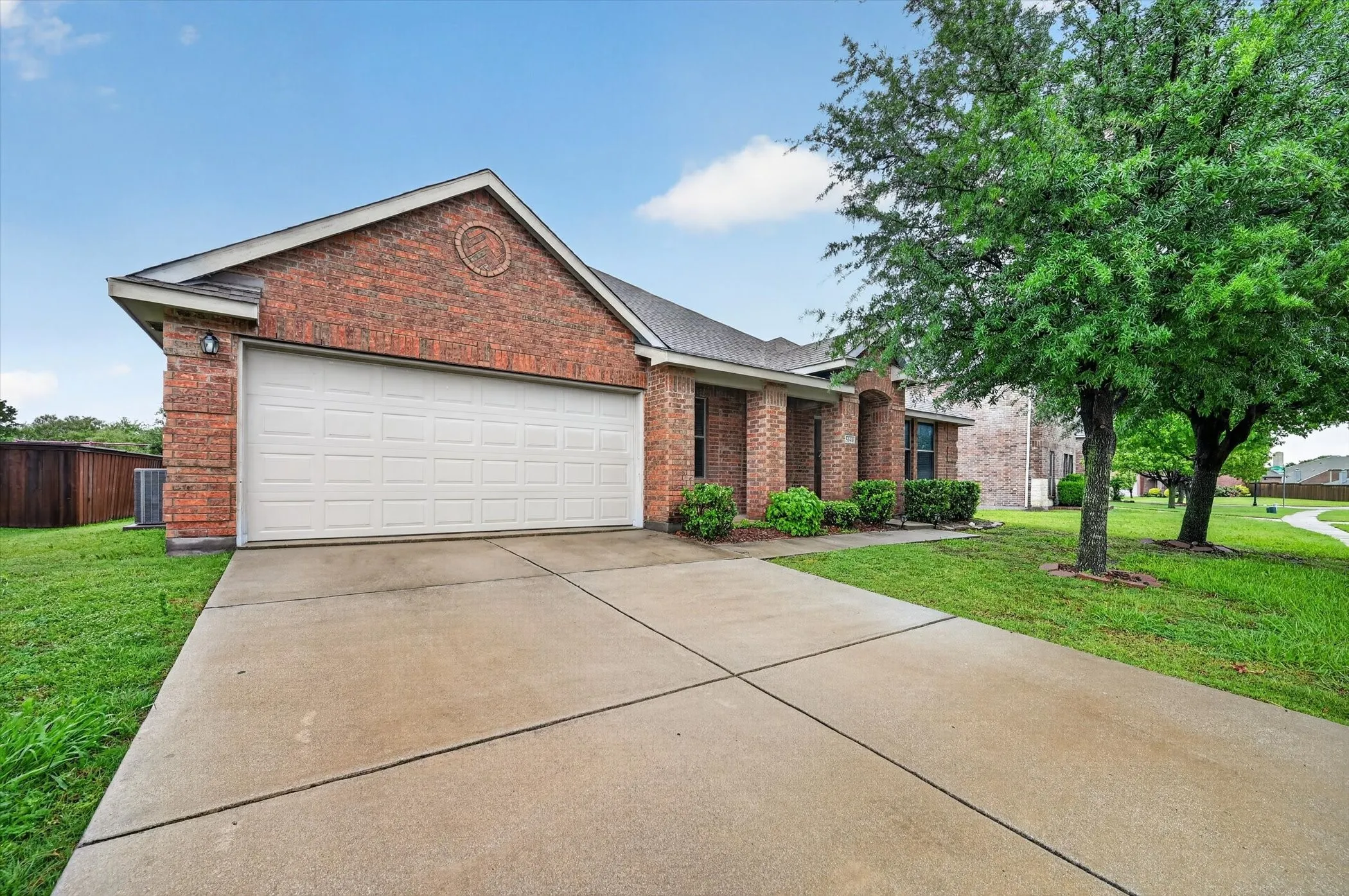 Brick facade, attached garage, concrete driveway, front landscaping, and mature trees