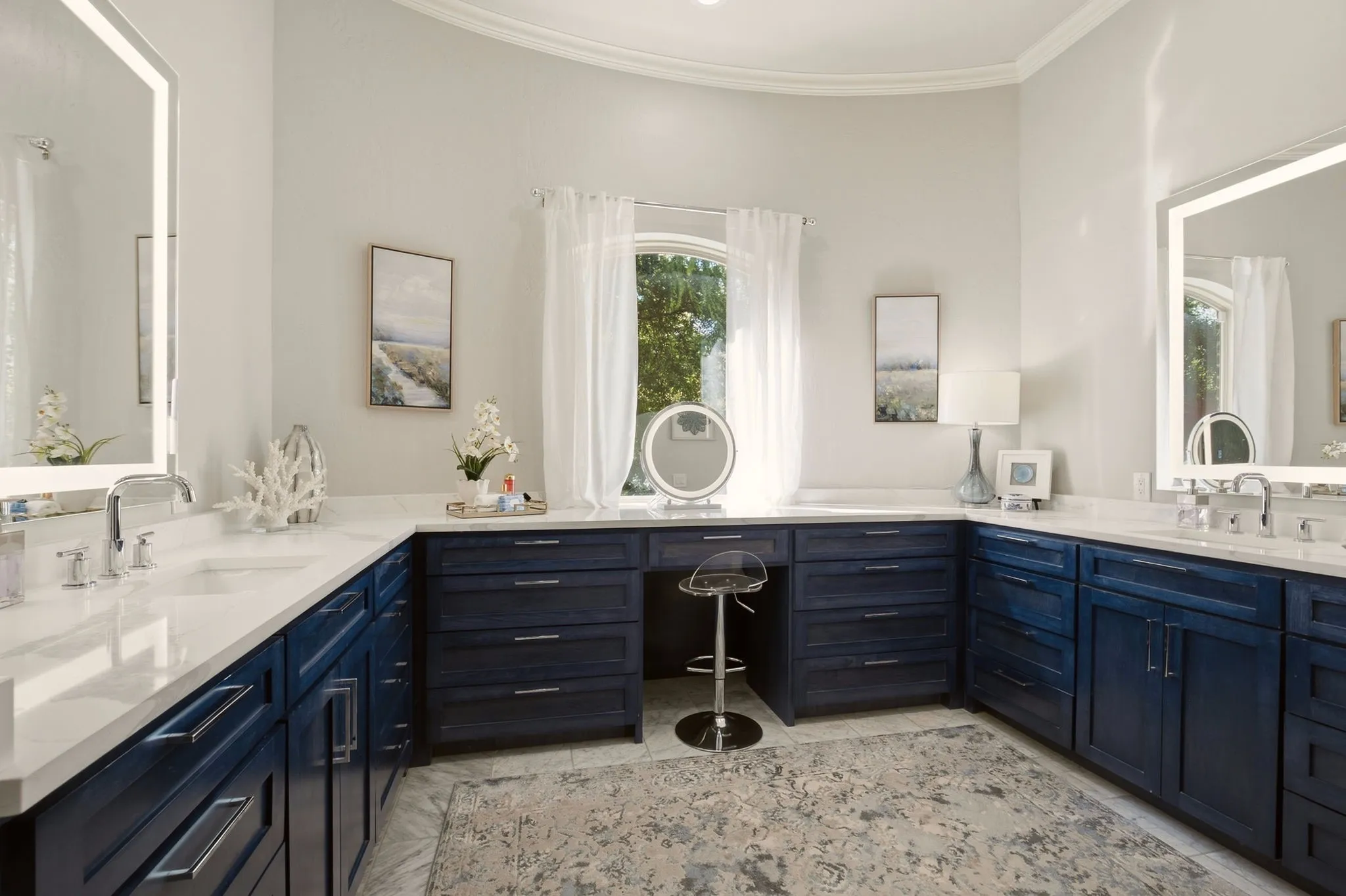 The luxurious resort-style primary bath offers lavish Carrera marble tile flooring and custom cabinetry that has been stained blue (not painted) accented by brushed-nickel hardware, offering generous deep-drawer storage for beauty and self-care essentials.