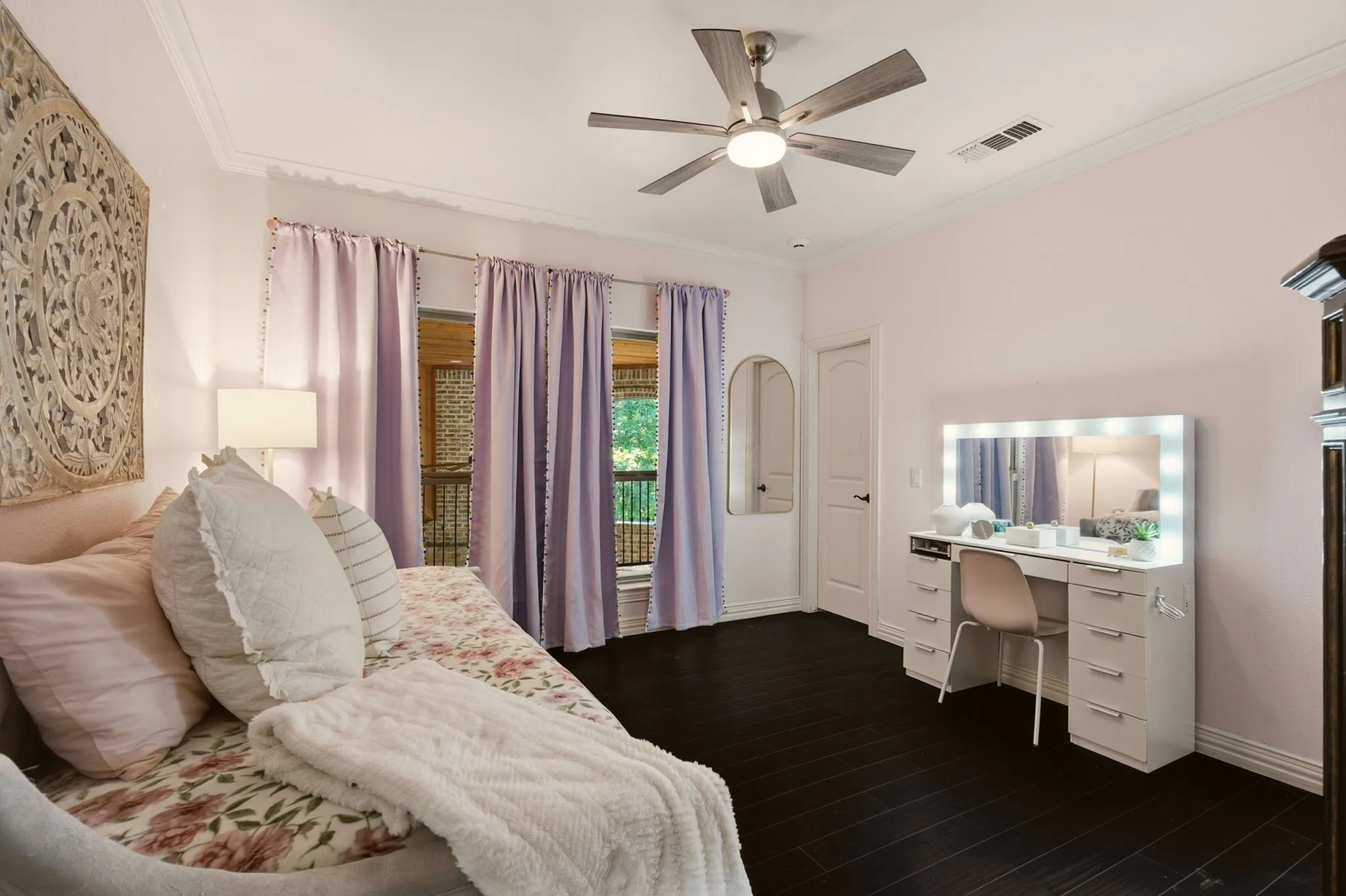 Bedroom 5 offers a cozy space with warm wood floors, crown molding, and a ceiling fan. Windows overlook the expansive second-floor terrace, creating a lovely connection to the home’s outdoor living spaces.