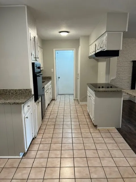 Galley kitchen featuring tile flooring, built-in wall oven, a cooktop on a central island, and white cabinetry with stone countertops