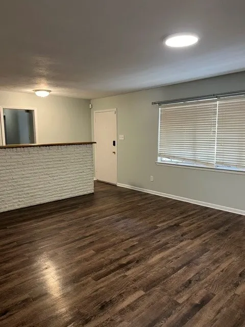 Wood-finish flooring throughout, white painted brick accent wall, white door with dark hardware, window with horizontal blinds, and ceiling-mounted lighting