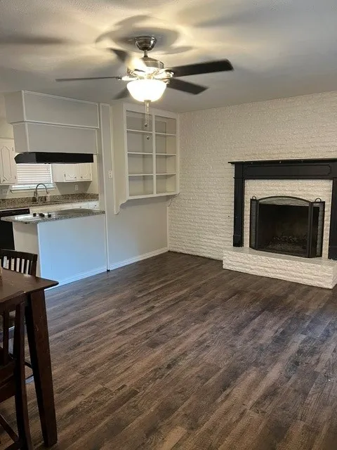 Wood-finish flooring, white brick fireplace with black mantel, built-in shelving, ceiling fan with light fixture, and kitchen with granite-finish countertops