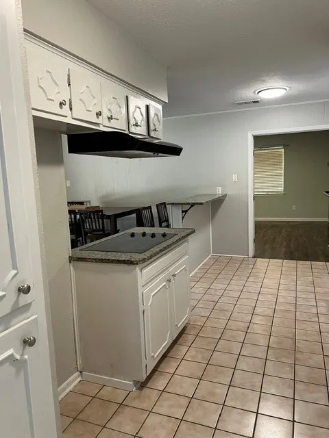 Kitchen featuring light-toned tile flooring, white cabinetry with decorative paneling, an integrated cooktop on a stone-finish countertop, and an overhead range hood