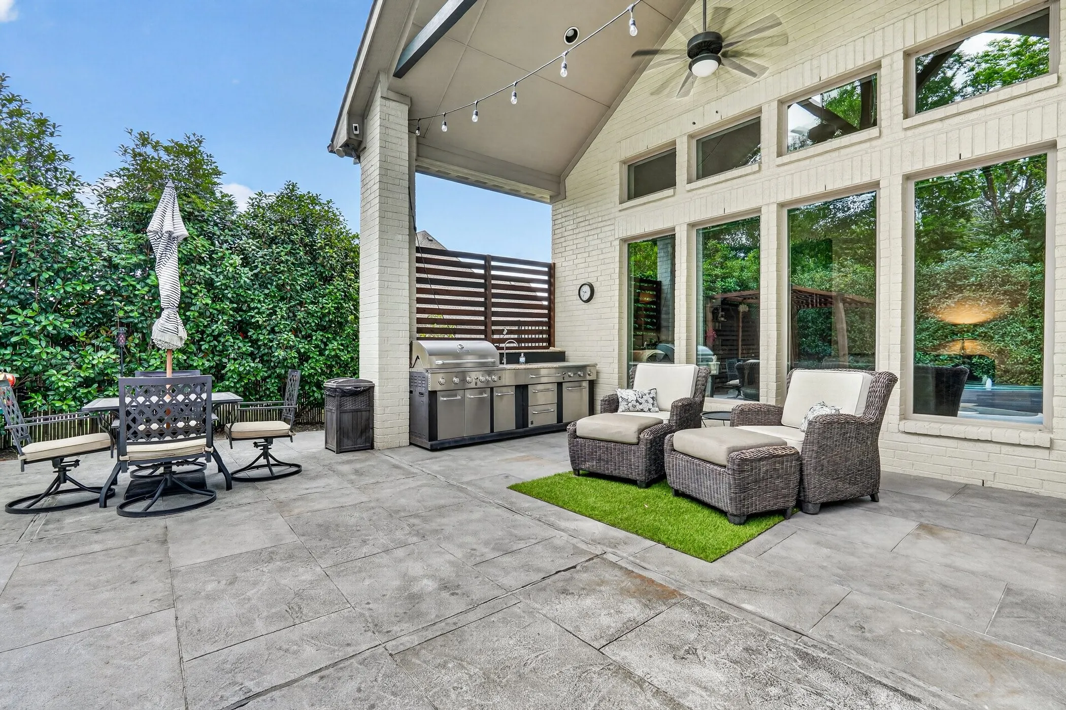 Expansive outdoor patio featuring large format pavers, a built-in outdoor kitchen with a stainless steel grill, a covered porch with a ceiling fan, string lighting, and floor-to-ceiling windows with transom elements