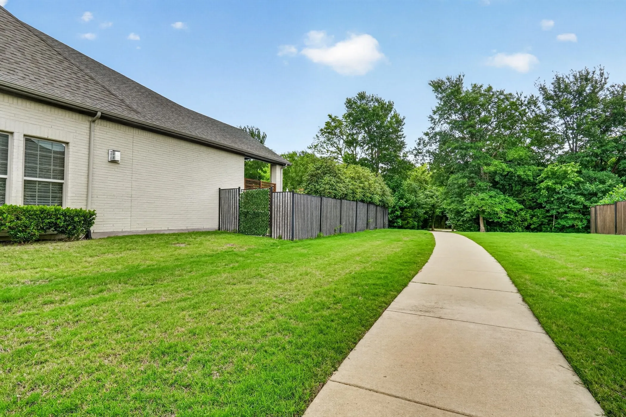Manicured lawn with a paved pathway, bordered by mature trees and a wood fence