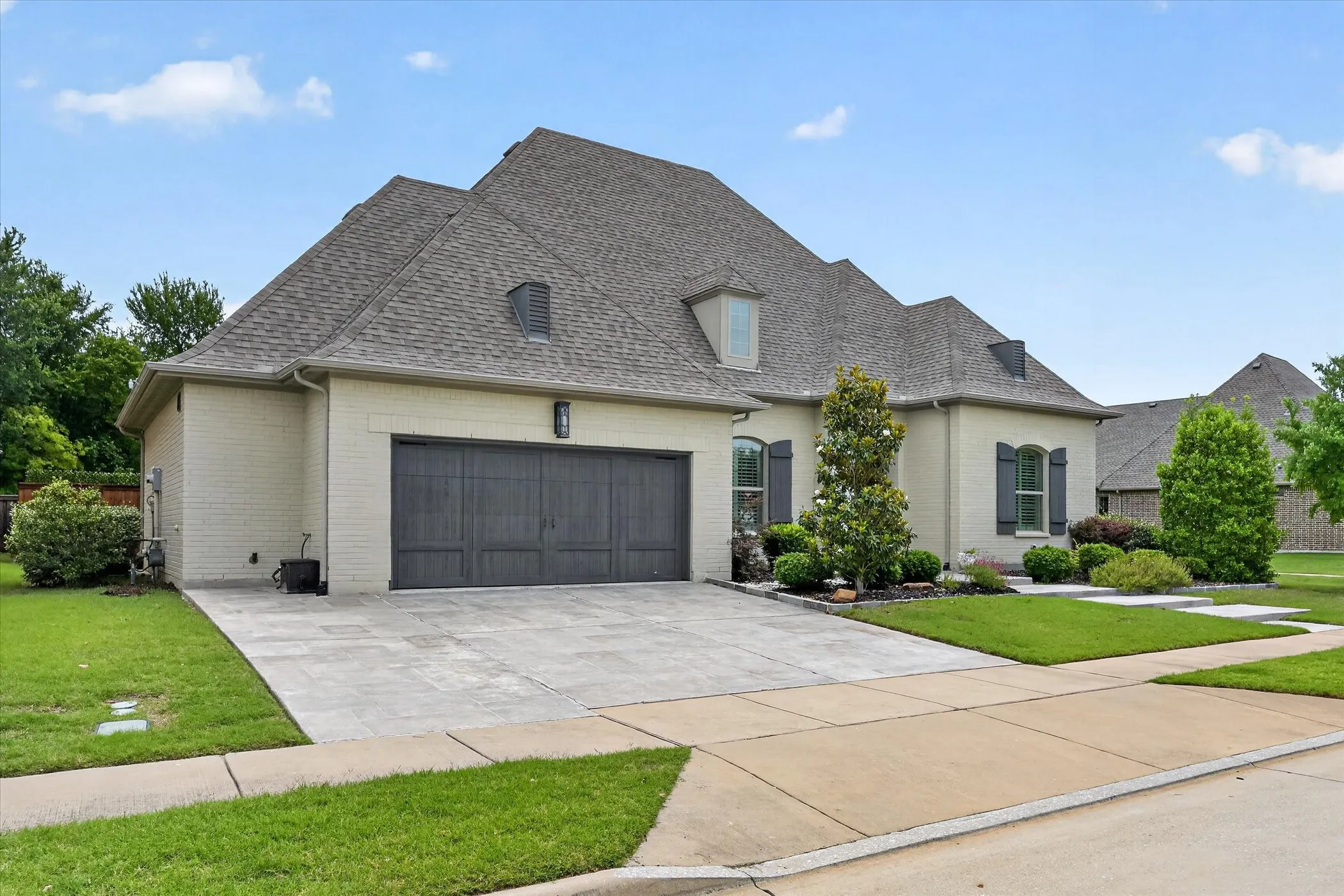 Cream brick exterior with dark shutters, dark garage door, and a gray shingle roof