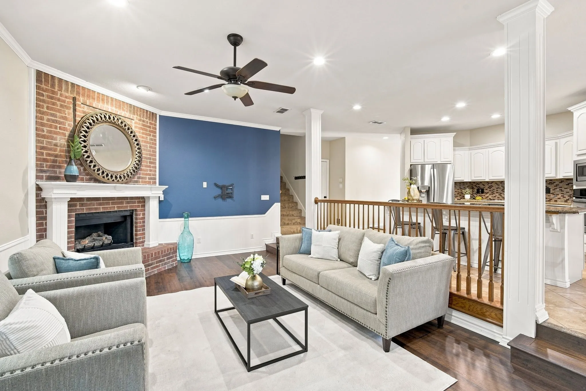 Sun-filled living area with gleaming floors, recessed lighting, a cozy brick fireplace, and an easy flow into the open white kitchen.