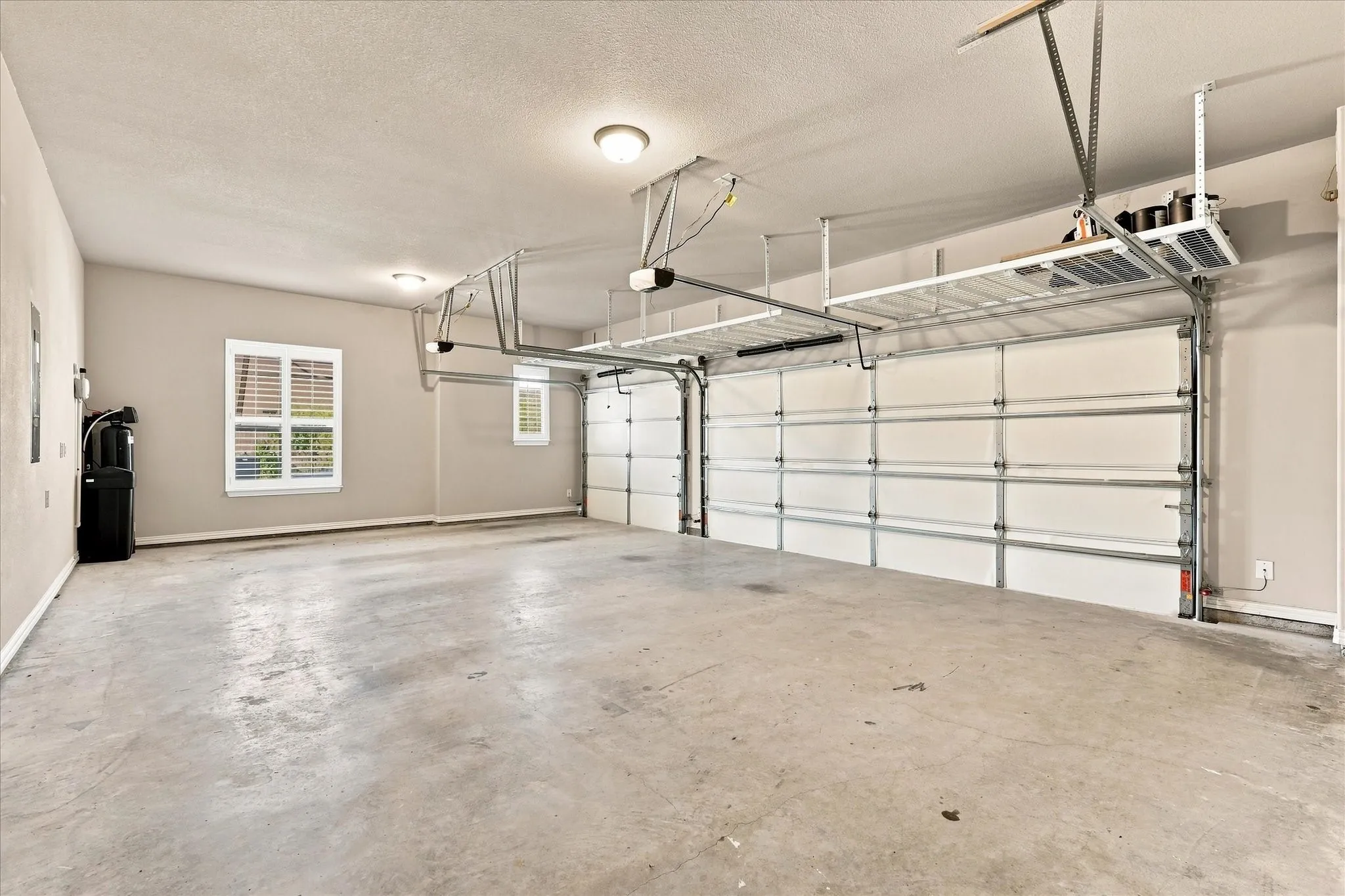 Two-car garage with concrete flooring, dual overhead garage doors, two windows with blinds, and built-in overhead storage racks
