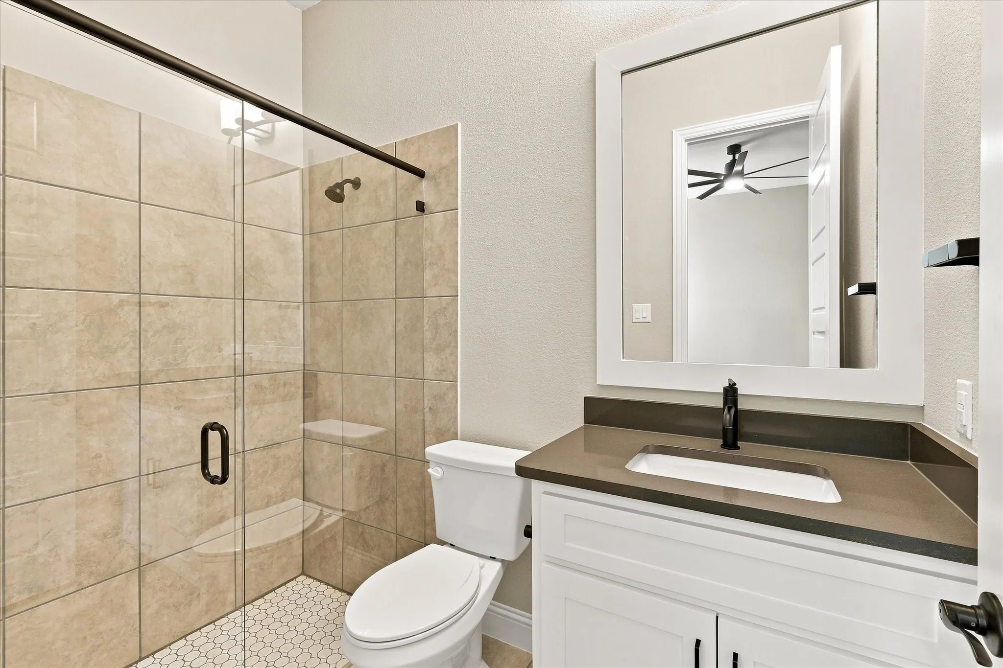 Glass-enclosed shower with tile surround and hexagonal tile flooring, white vanity with integrated sink and dark countertop, and framed vanity mirror