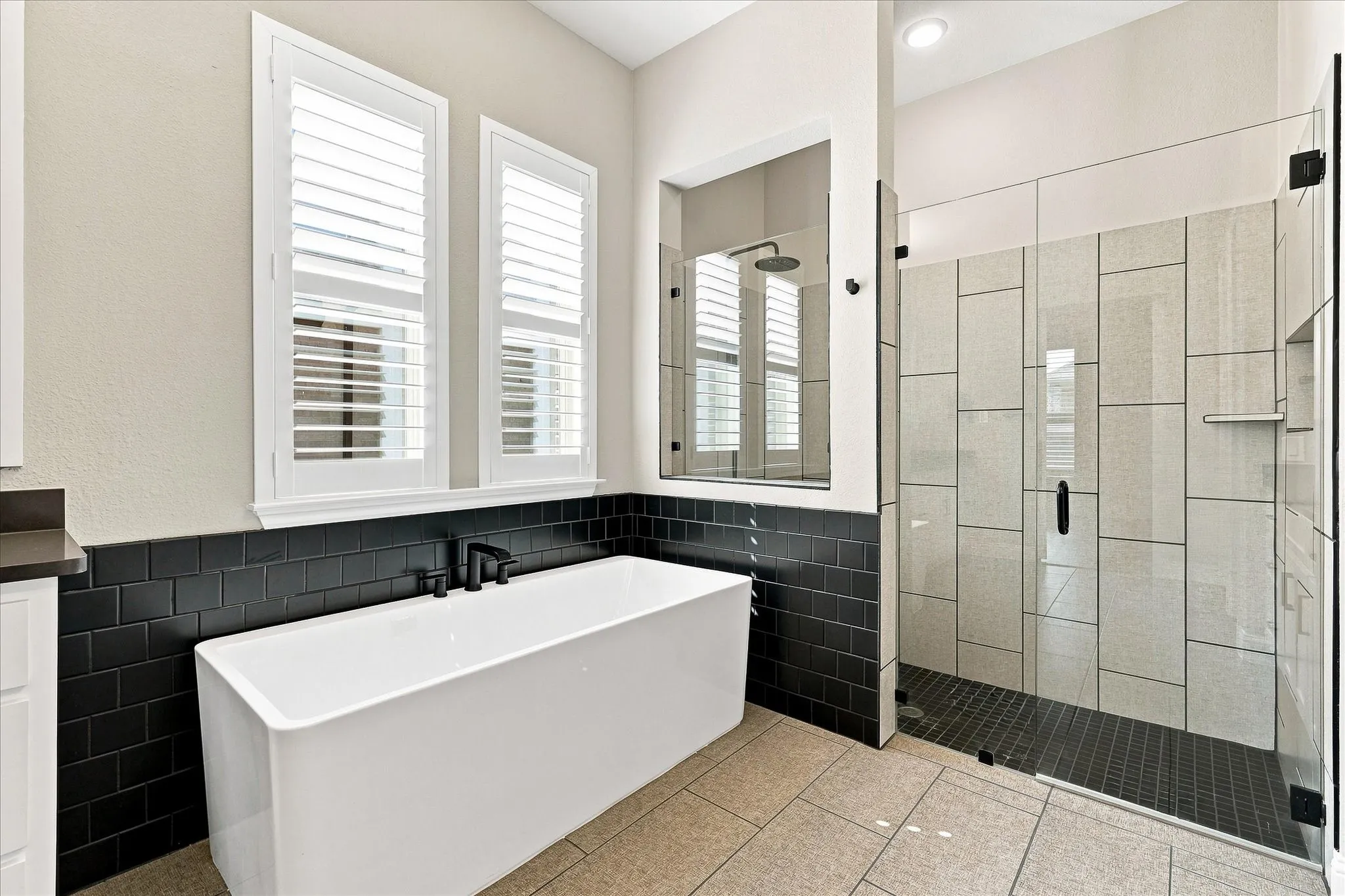 Freestanding soaking tub with matte black fixtures, framed by a black subway tile accent wall and plantation-style window shutters