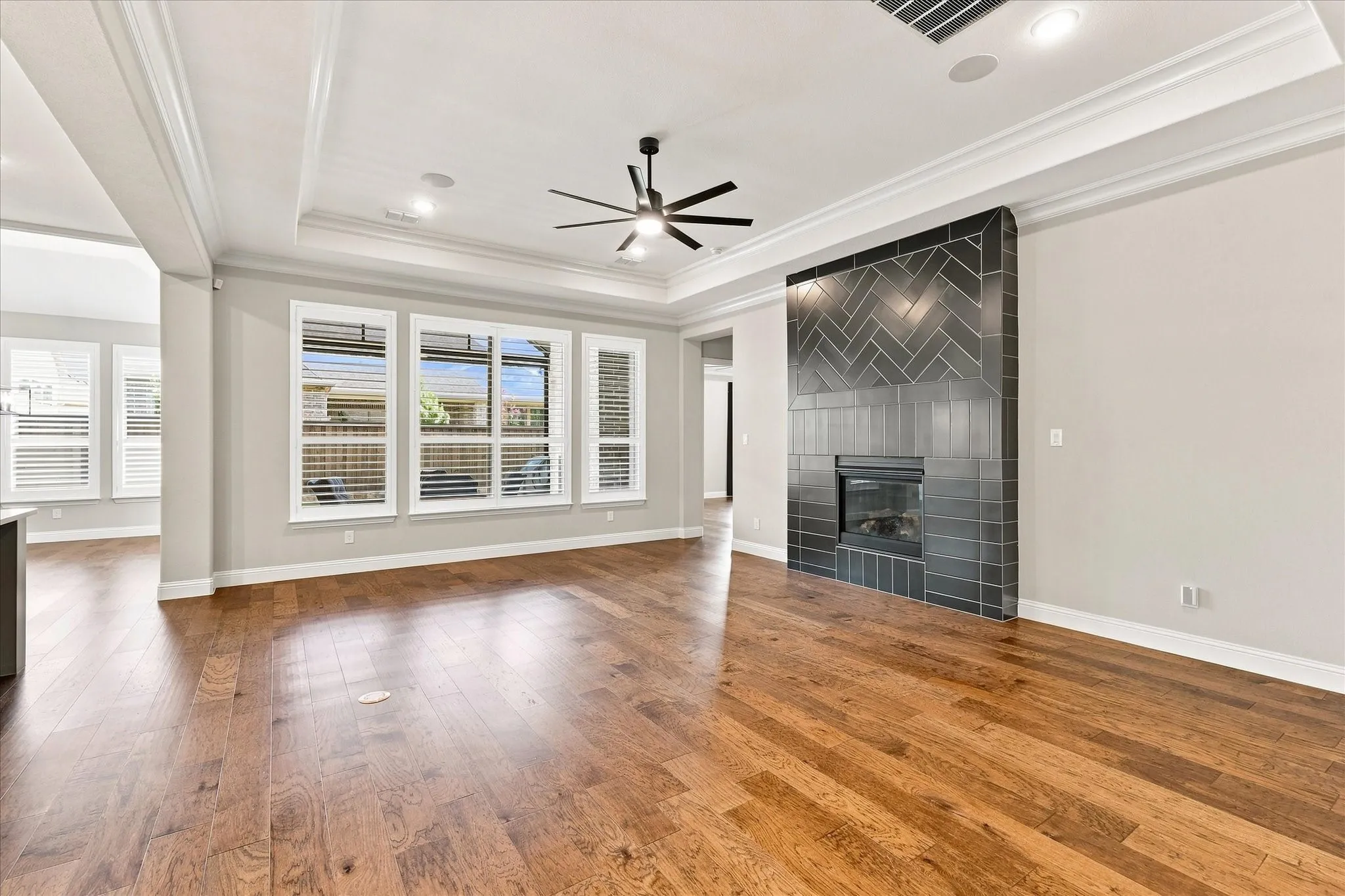 Wood-finish flooring, tray ceiling, recessed lighting, ceiling fan, and a fireplace with a herringbone tile surround