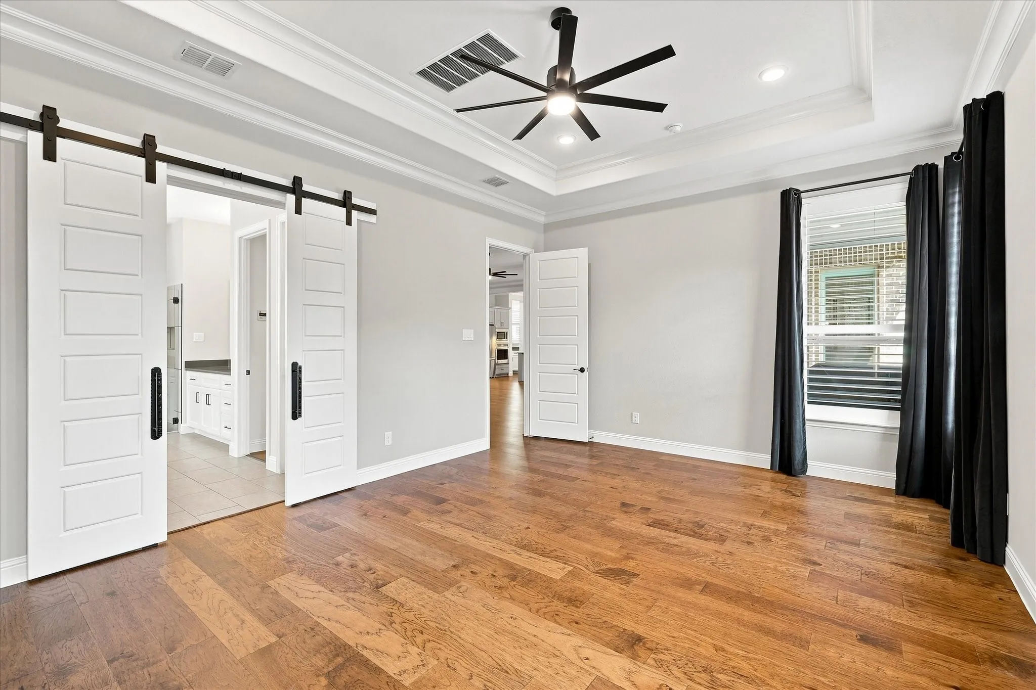 Wood-finish flooring, tray ceiling, ceiling fan, and white barn doors with matte black hardware