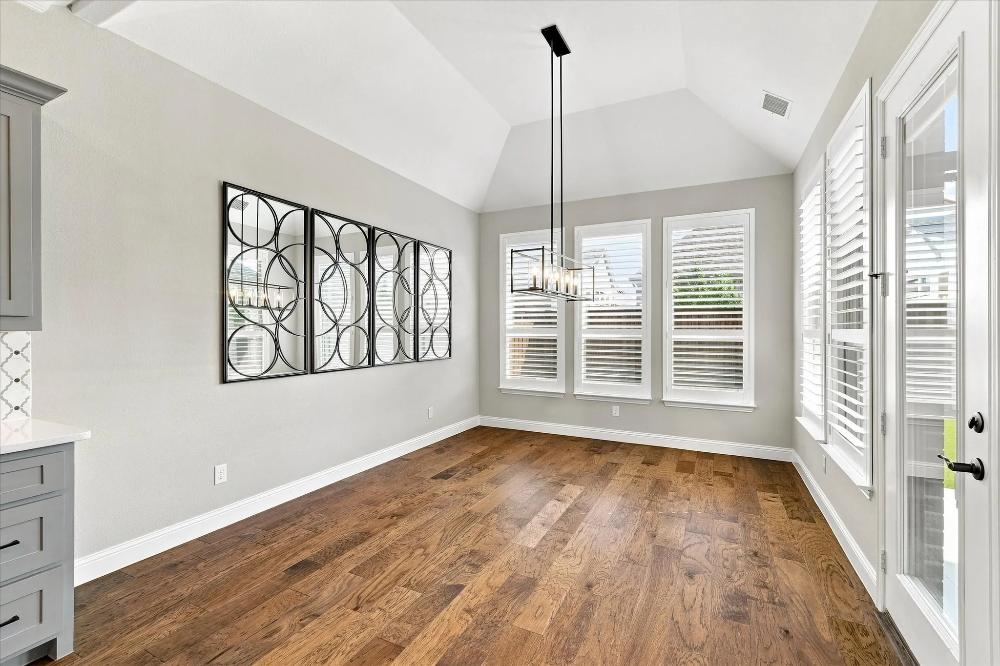 Vaulted ceiling, wood-finish flooring, multiple windows with plantation shutters, overhead chandelier, and a glass-paneled door