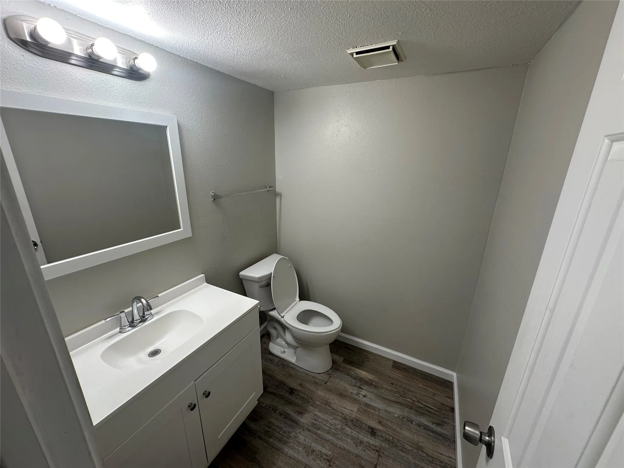 White vanity with integrated sink, chrome faucet, wood-finish flooring, wall-mounted mirror, and overhead vanity lighting