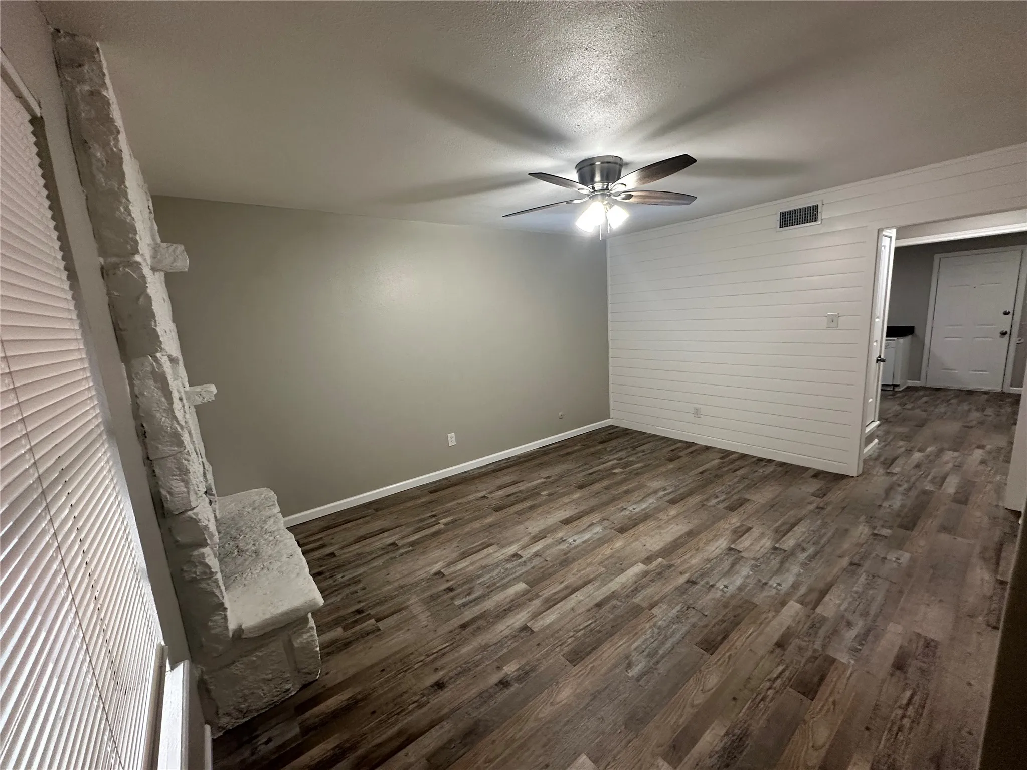 Wood-finish flooring throughout, stone fireplace with mantel, shiplap accent wall, and ceiling fan with integrated lighting